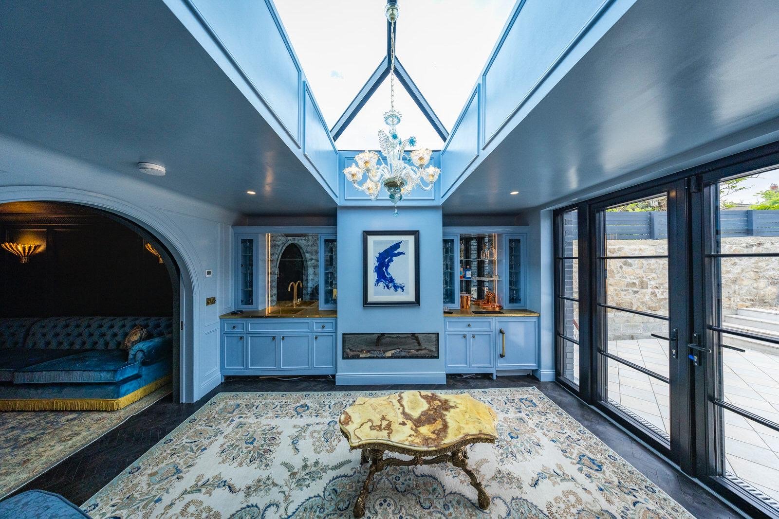 Interior view of a luxurious lounge that has lacquered walls and ceiling, a pyramid skylight, antique murano glass chandelier, framed artwork, built-in cabinets with brass accents, a fireplace, antique persian rug, and a glass door leading outside.