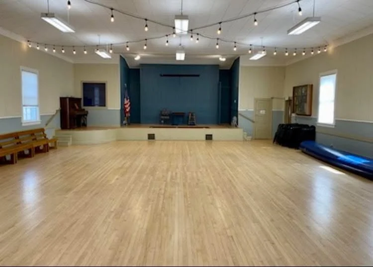 Empty community hall with a small stage, wooden floor, string lights, and benches along the walls.