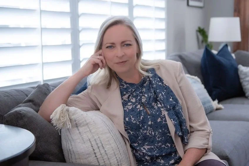 A woman with blonde hair sitting on a gray couch, leaning on a pillow, with her hand resting on her face, looking at the camera in a well-lit living room with white blinds and cushions.