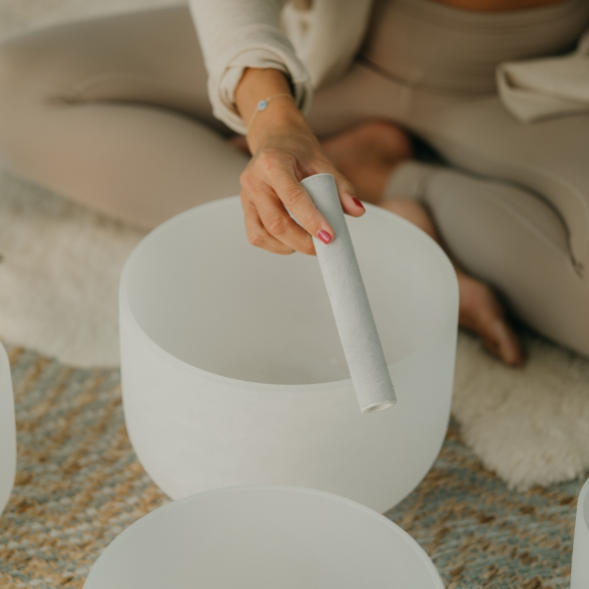 A person holding a white mallet near a white singing bowl, sitting on a beige sofa with a patterned rug underneath.