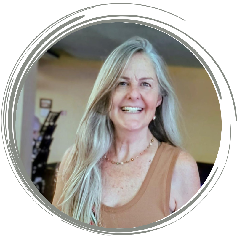 A smiling woman with long gray hair, wearing a beige sleeveless top and a gold necklace, indoors with a blurred background.