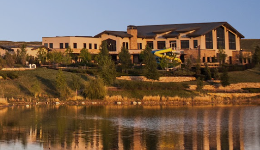Large house or clubhouse near a body of water, with trees and landscaping around, under a clear sky.