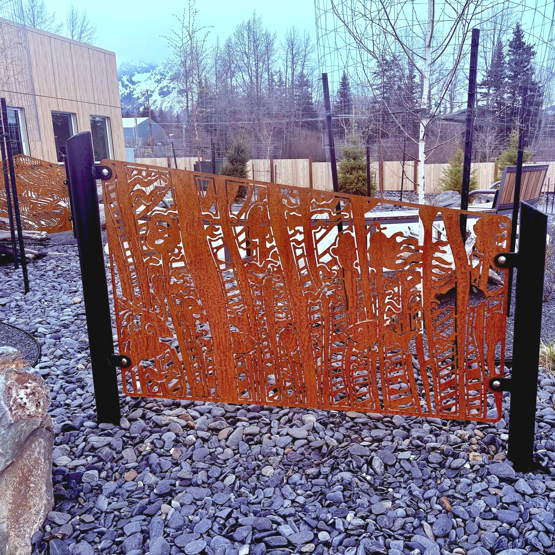 Decorative metal gate with mountain and forest design, set in a garden with rocks, trees, and a modern building in the background.
