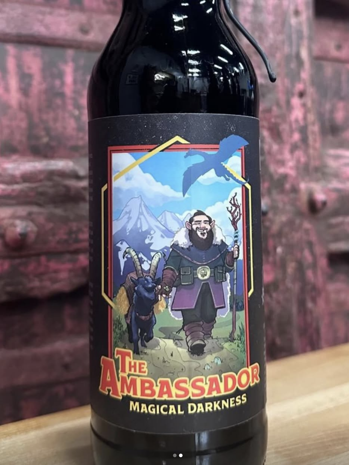 A dark bottle of beer with a colorful label featuring a cartoon wizard with a beard, holding a staff and a goat standing beside him. The background includes mountains, clouds, and a flying dragon. The label reads 'The Ambassador Magical Darkness' in bold letters.