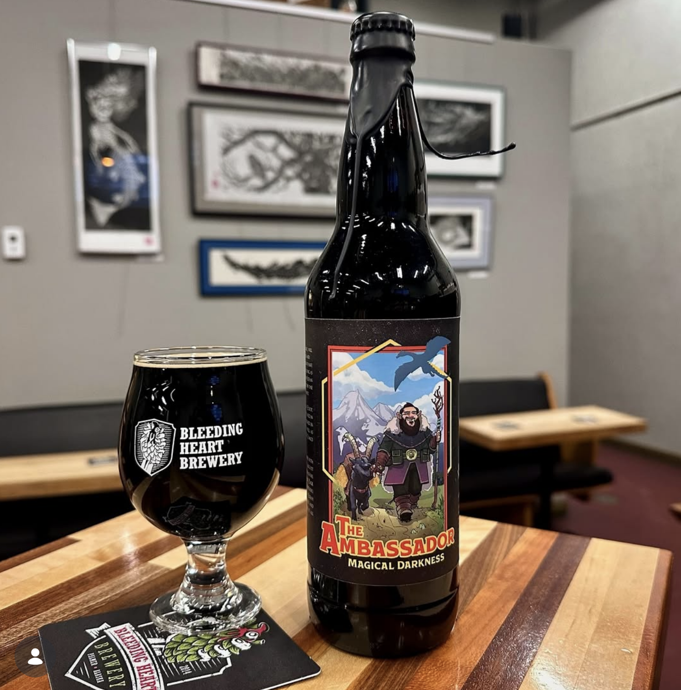 A dark-colored beer bottle labeled 'The Ambassador: Magical Darkness' from Bleeding Heart Brewery, with a matching dark beer in a glass, on a wooden table in a brewery or pub setting with framed artwork on the wall in the background.