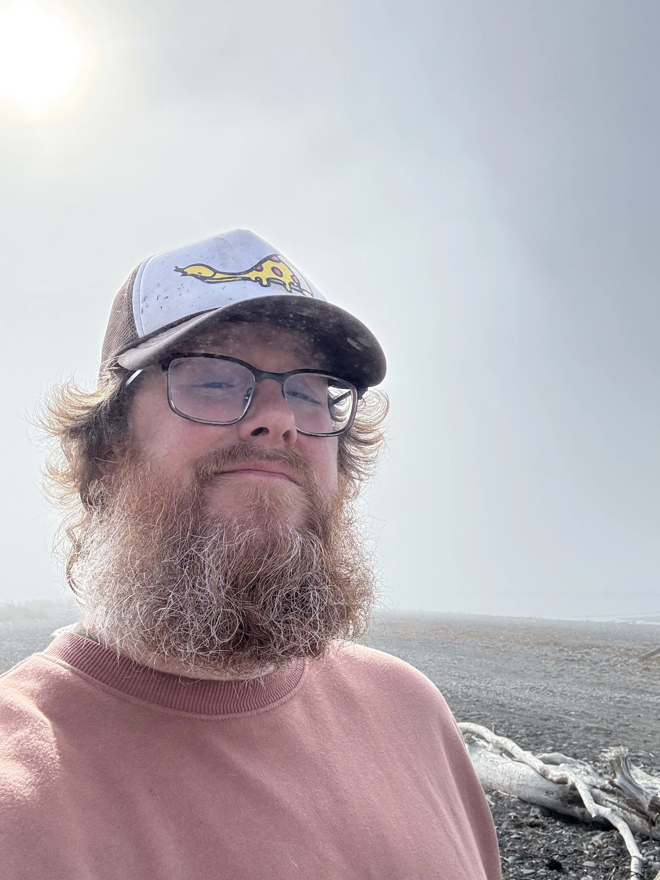 A man with a beard and glasses wearing a baseball cap and pink sweatshirt, standing outdoors in a foggy landscape with a log on the ground.