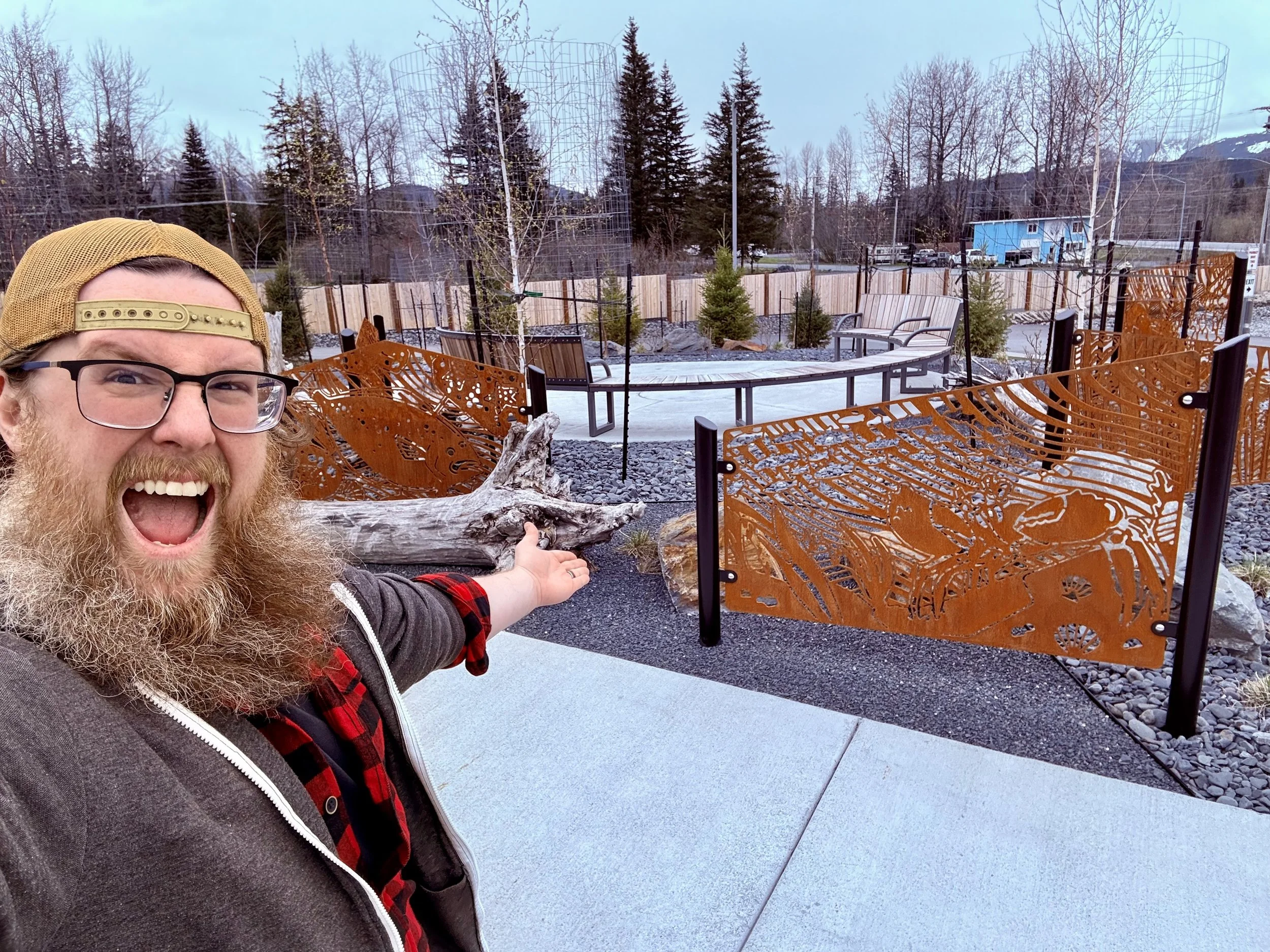 A man with glasses, a beard, and a baseball cap is smiling and pointing at an outdoor art installation with rust-colored metal panels depicting various animals. The background includes trees, a wooden fence, and a small building.