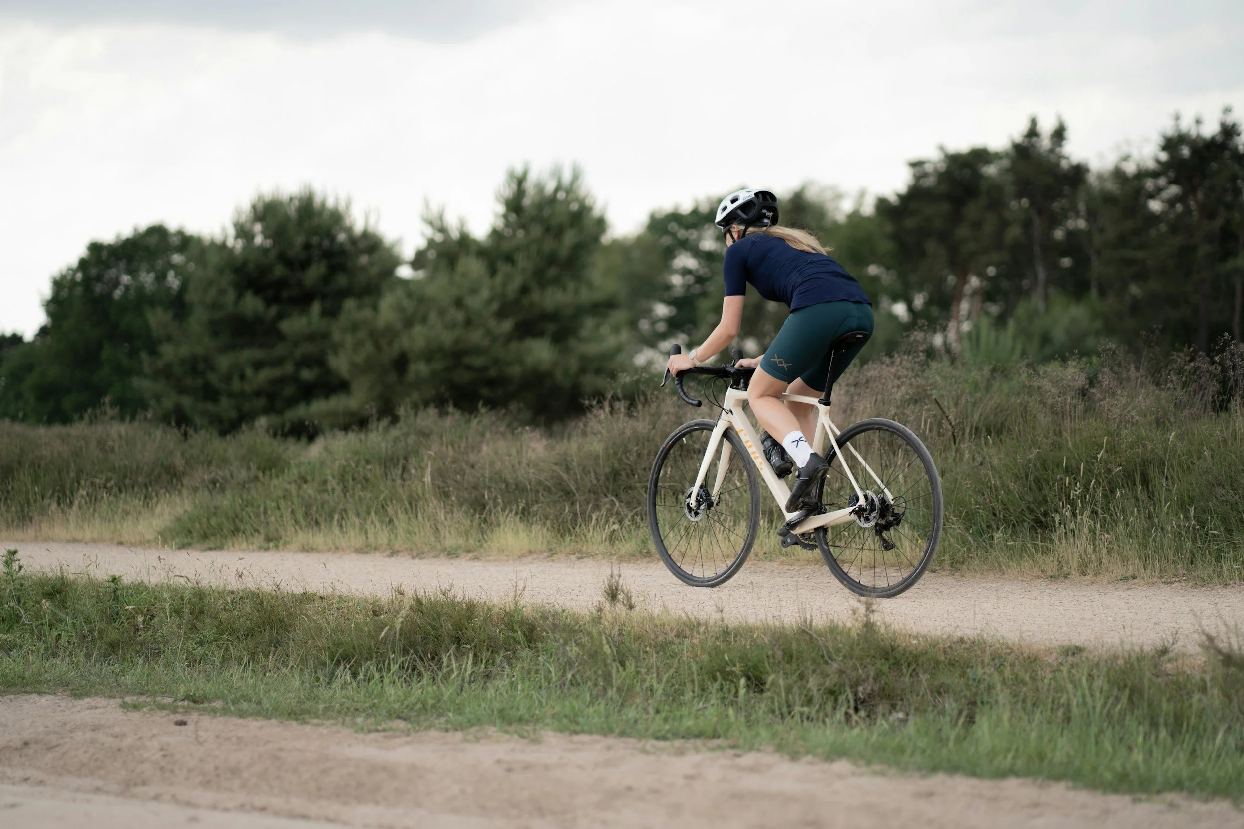 A woman riding a white bicycle on a dirt path through a green landscape with trees.