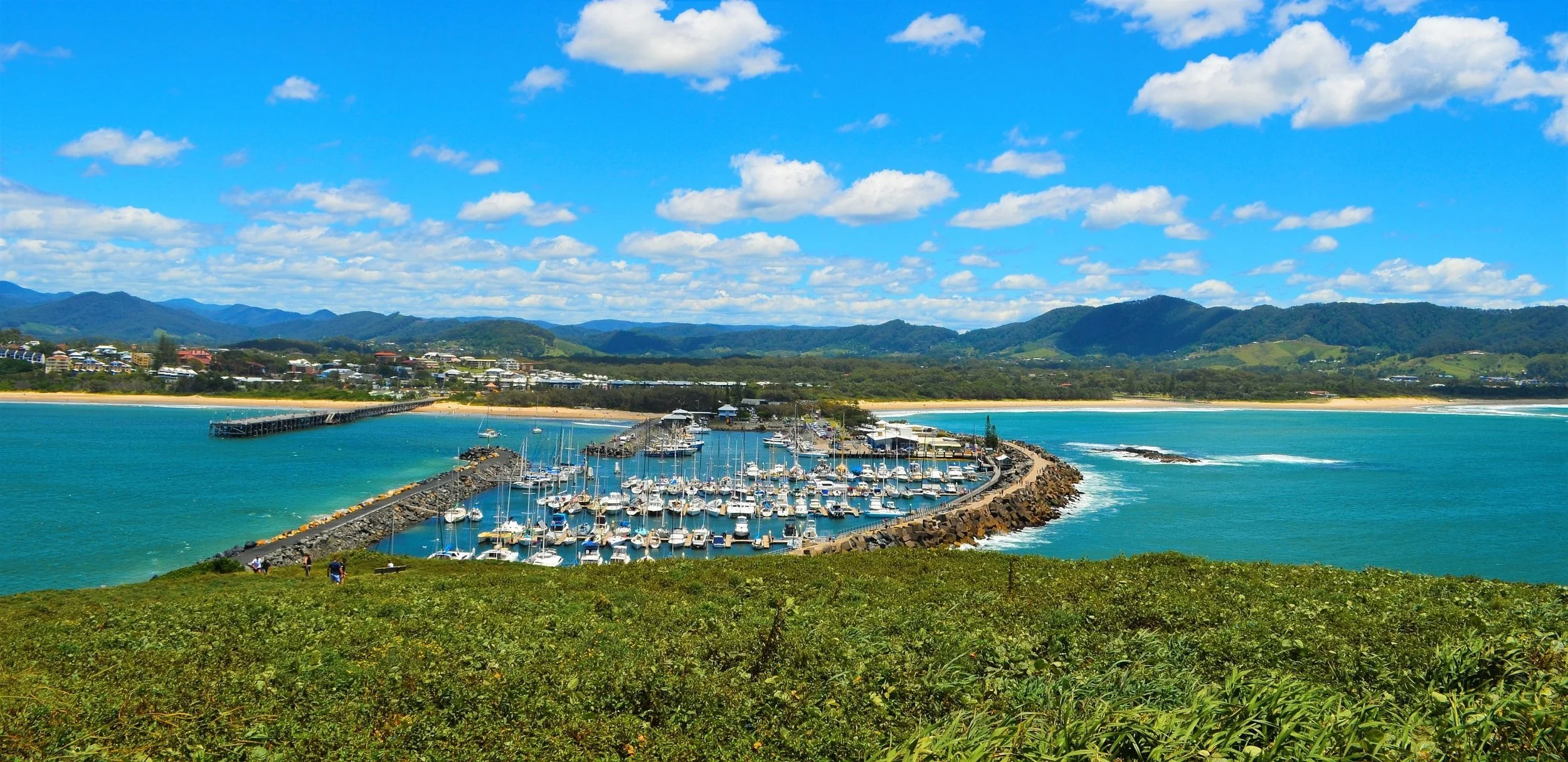 A scenic view of a marina with numerous boats docked, surrounded by blue water, with a coastal town, green hills, and mountains in the background under a partly cloudy sky.