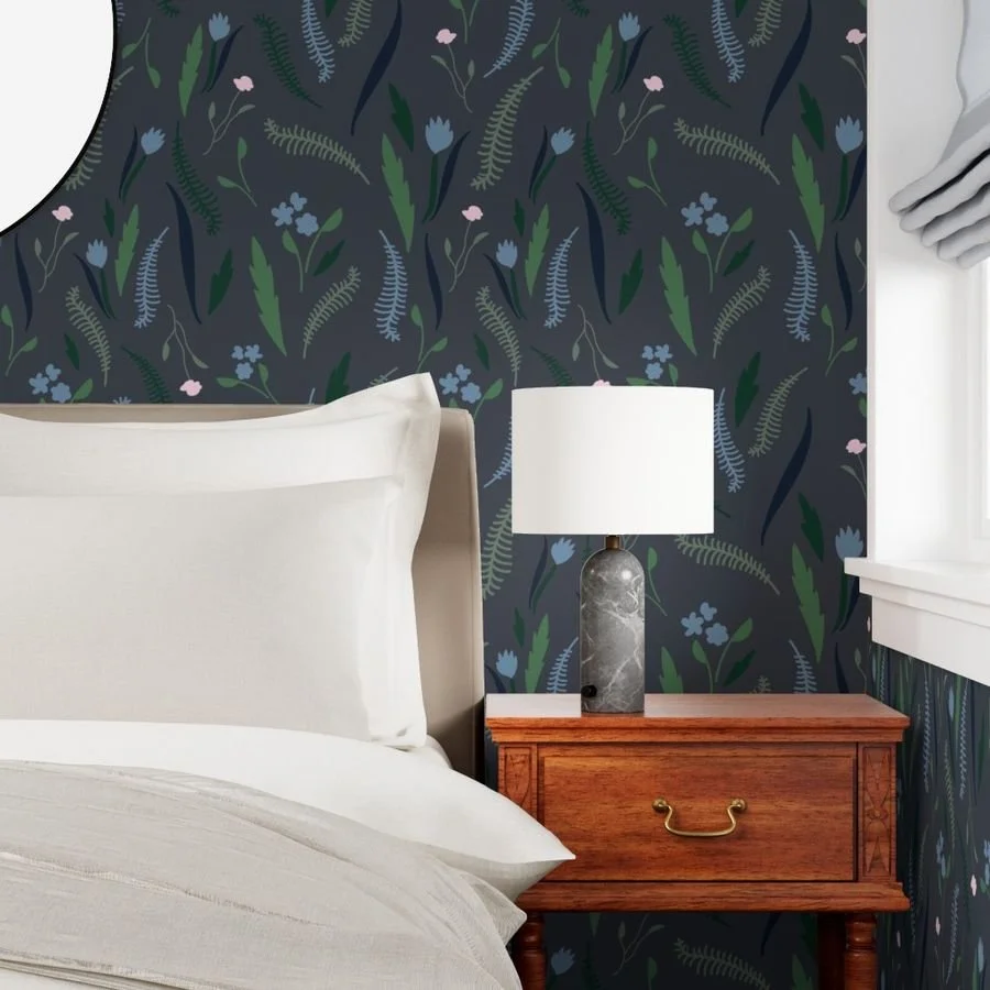 A bedroom corner featuring a bed with white bedding, a wooden nightstand, a lamp with a white shade, and dark moody floral wallpaper in shades of blue, green, and charcoal gray.