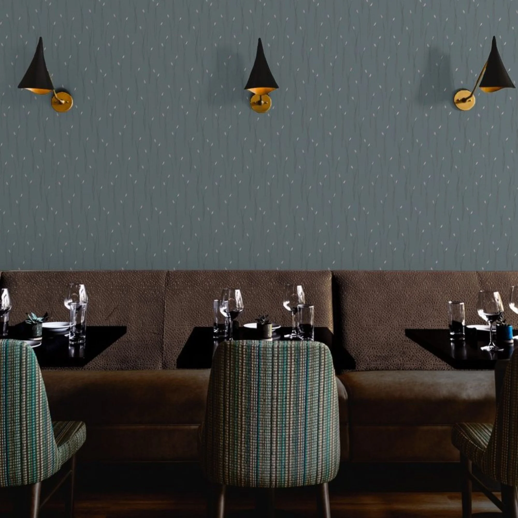 Empty restaurant with tables set for dining, chairs with plaid fabric covers, wine glasses, and small potted plants, and a decorative deep teal wallpaper behind, featuring tall reed grasses.