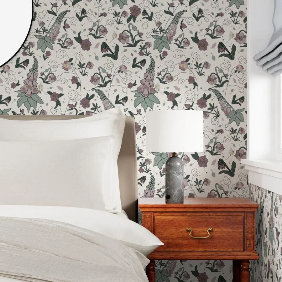 A bedroom with a maximalist, green, pink and cream floral wallpaper, a white headboard, a nightstand with a lamp and white lampshade, white pillow and bedsheet, and a window with a white curtain.
