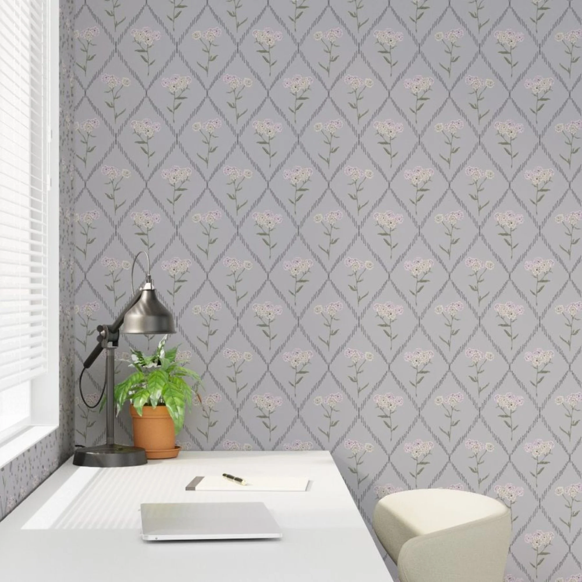 A small white table with a pen and notebook, a potted plant on a stand, a desk lamp, and a chair, with dove gray floral patterned wallpaper and a window with blinds in the background.