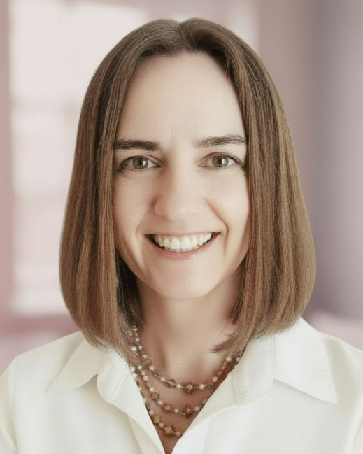 A woman with shoulder-length brown hair smiling at the camera, wearing a white collared shirt and a layered pearl necklace. The background is blurred with neutral tones.