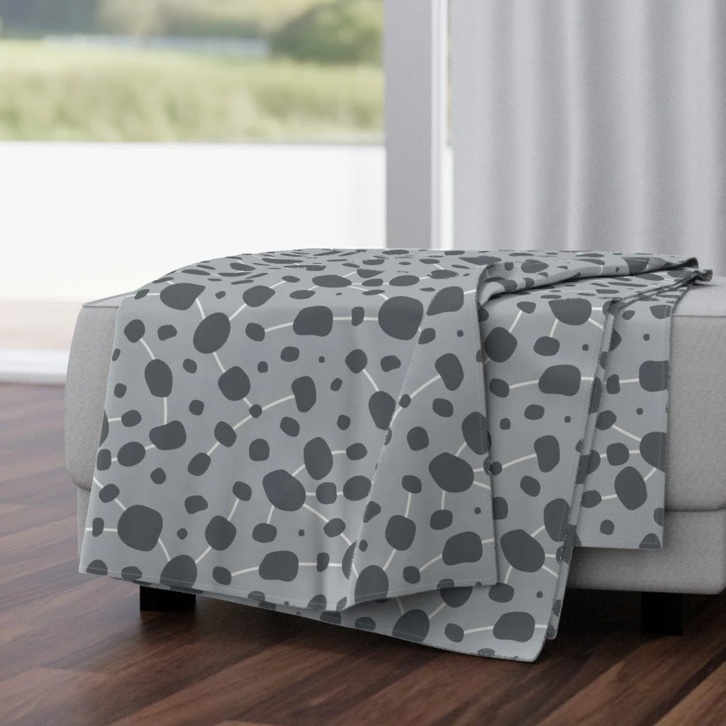 Gray blanket with black abstract spots draped over a light-colored sofa in a modern living room with large windows.