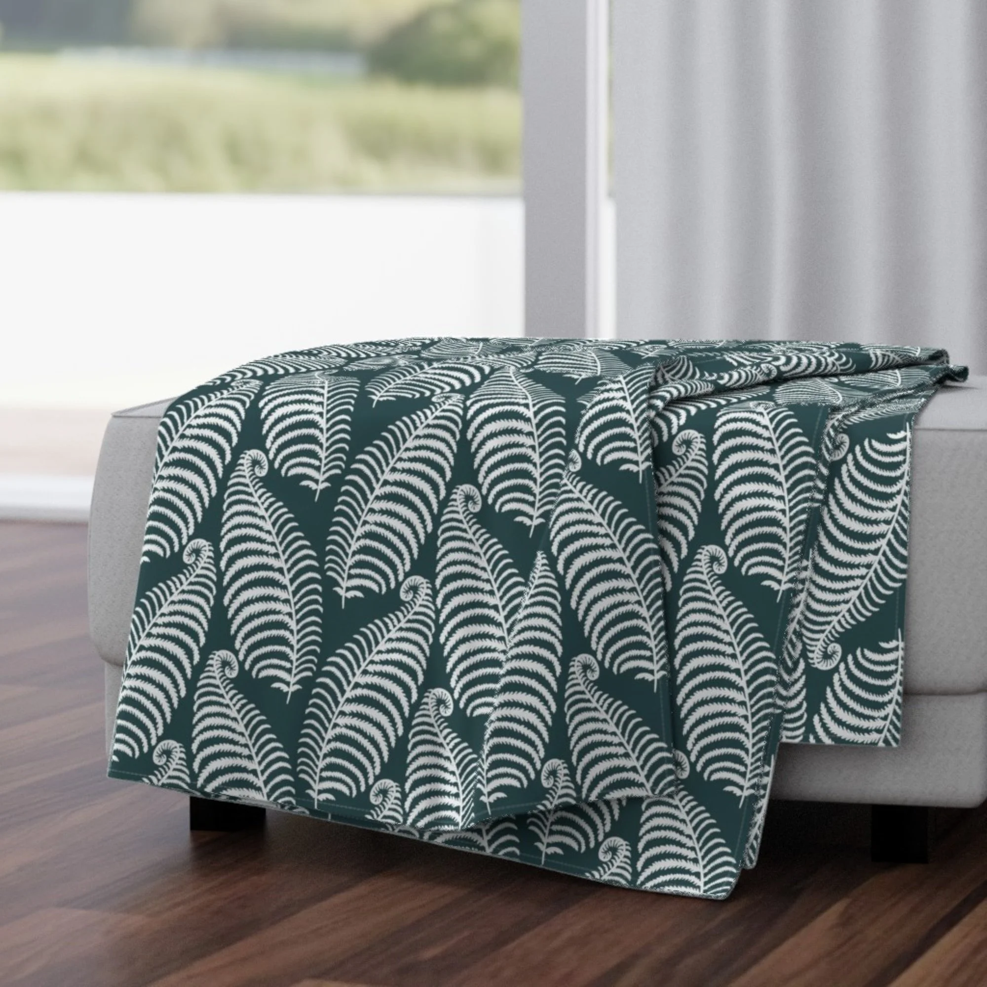 A decorative throw blanket with a dark teal and white fern pattern draped over a white table, placed on a dark wooden floor near a window with a blurred green outdoor landscape visible.