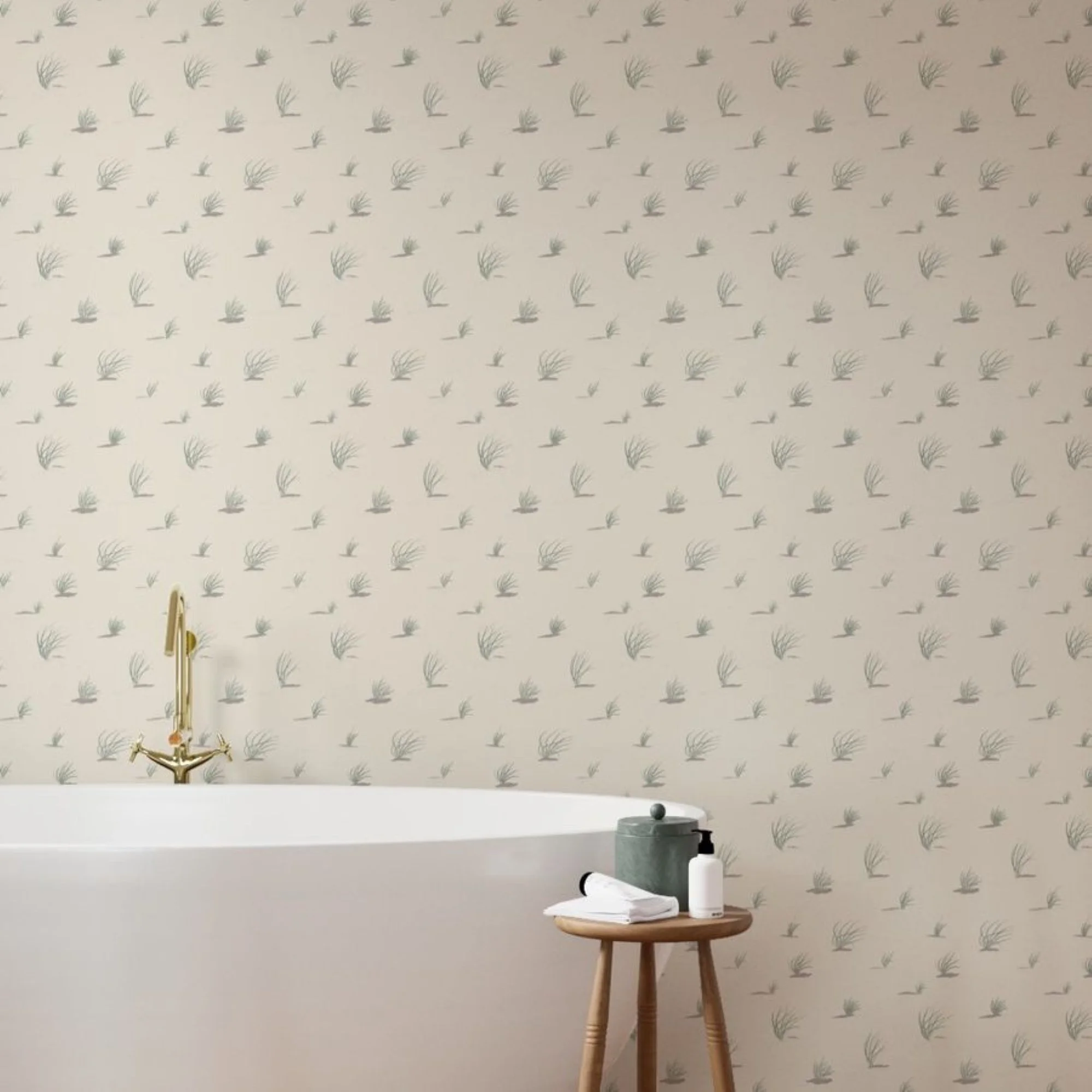 Interior room with wallpaper featuring small, light teal green dune grass botanical illustrations, a small wooden table with books, a black and a white water bottle, and a gold faucet and white tub.
