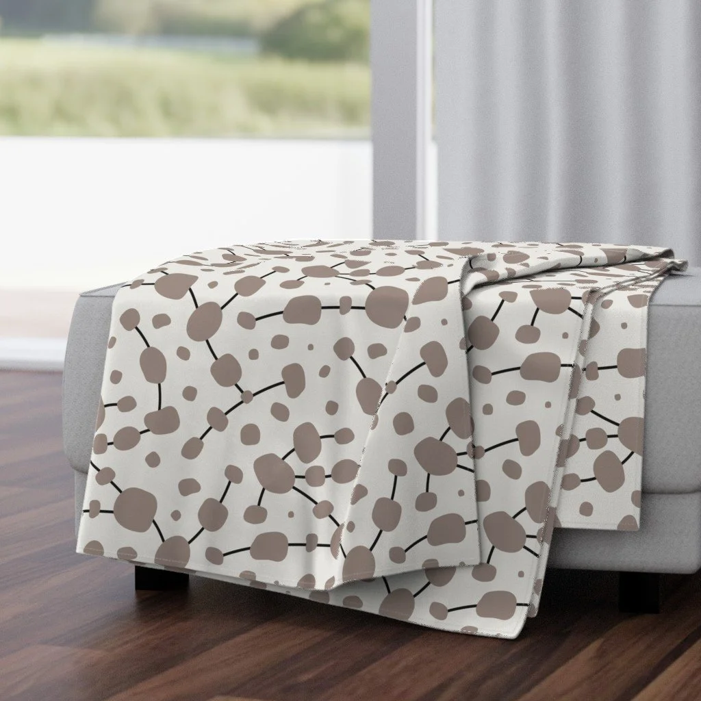 A blanket with a cream and brown abstract dot pattern is draped over the side of a light gray upholstered sofa in a modern living room with hardwood floors and a large window in the background.