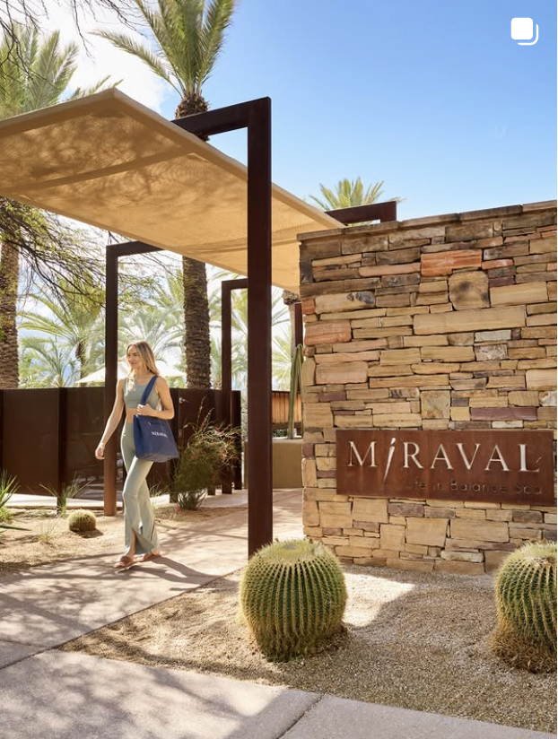 A woman walking through a desert-like area with cactus plants, palm trees, and a stone wall with a sign that reads 'MIRAVAL wellness & balance spa' under a shade canopy.