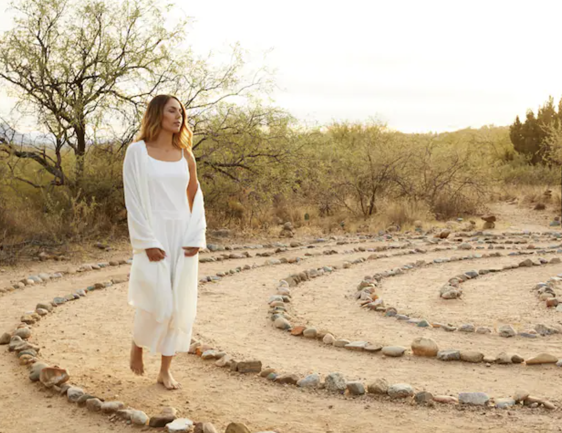 A woman with shoulder-length wavy hair, dressed in a flowy white outfit, walking barefoot on a dirt path lined with stones in a desert landscape during sunset, with sparse trees and distant hills.