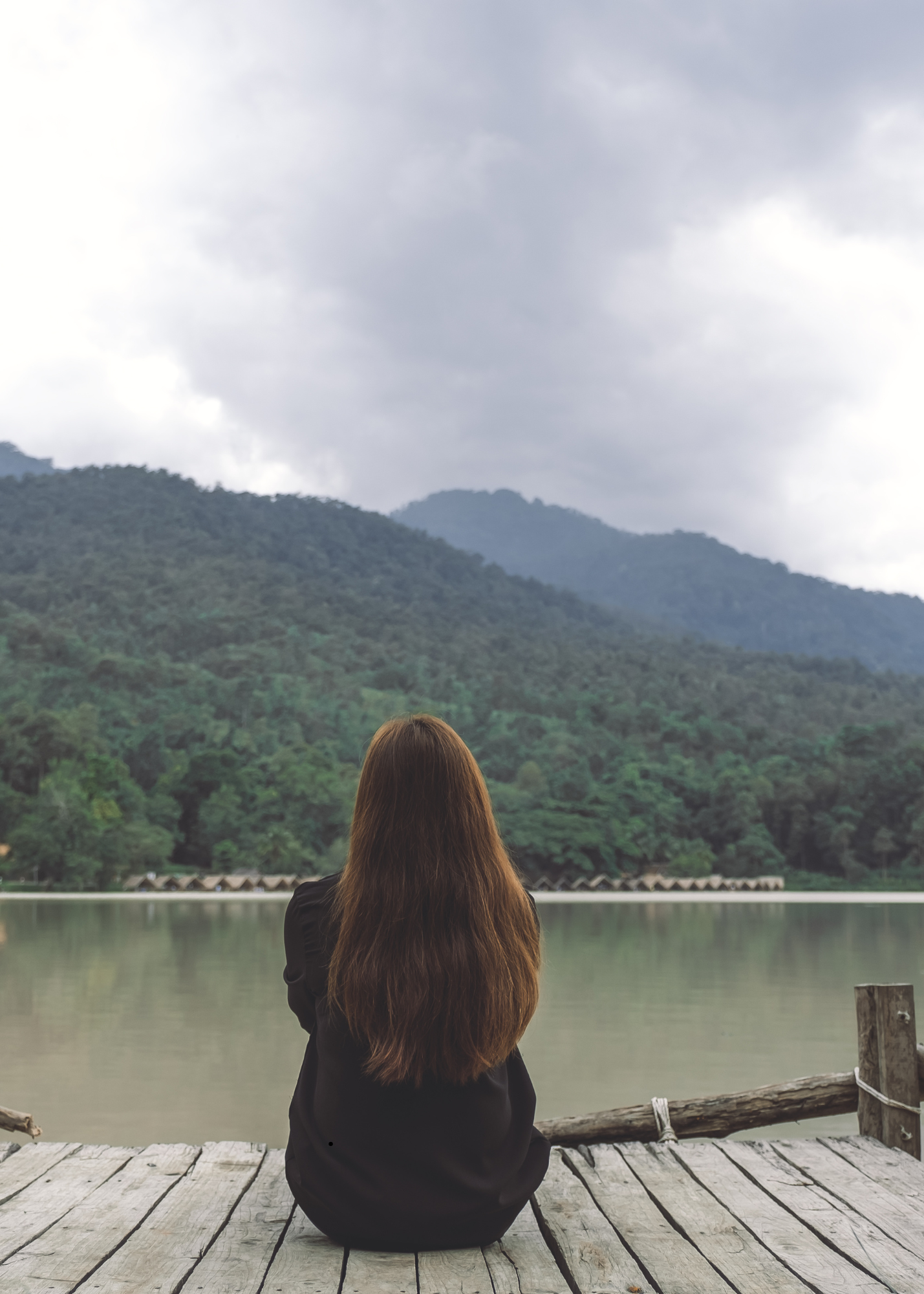 A woman with long brown hair sitting cross-legged on a wooden dock, facing a lake with mountains in the background and cloudy skies.
