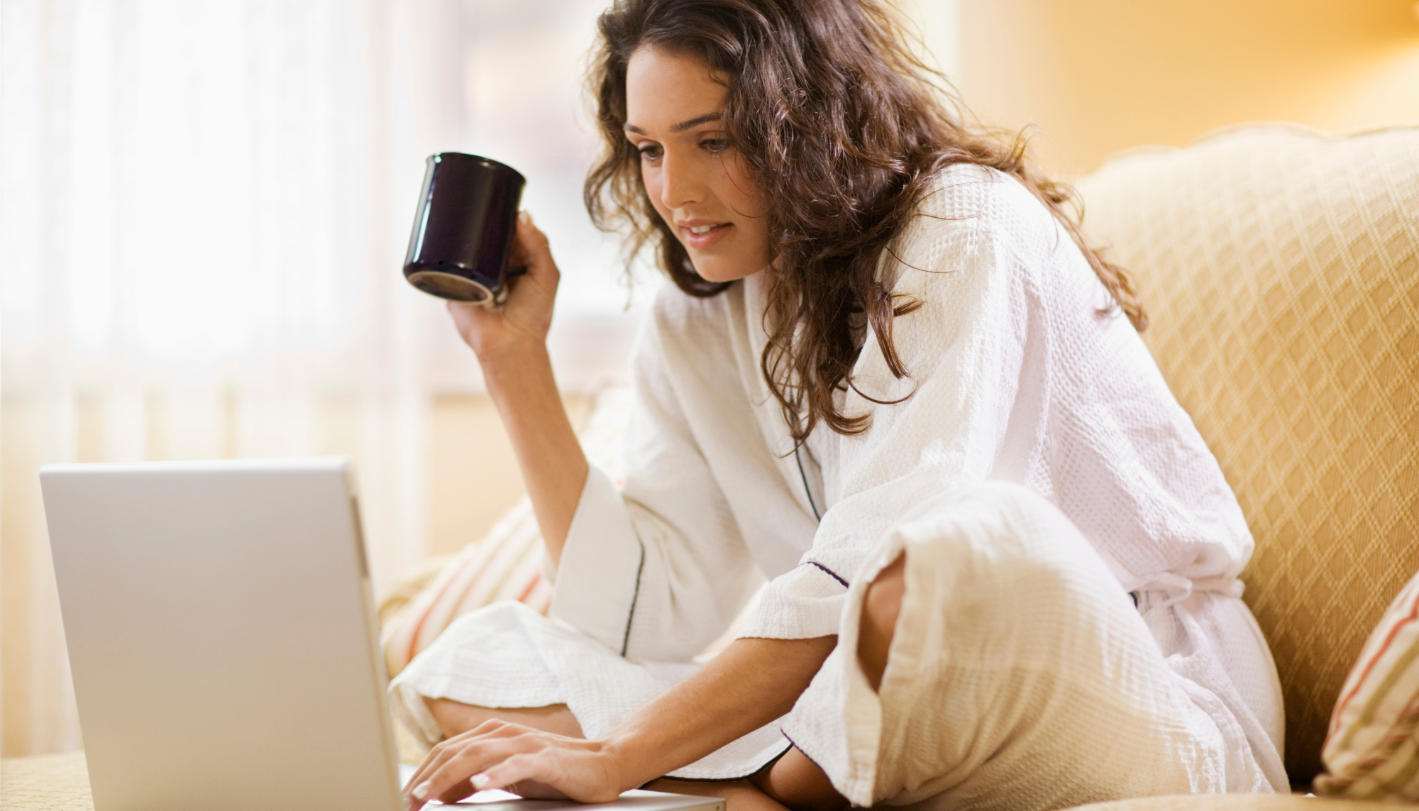 Woman sitting on a beige couch holding a black mug, wearing a white textured robe, with a laptop in front of her, in a well-lit room.