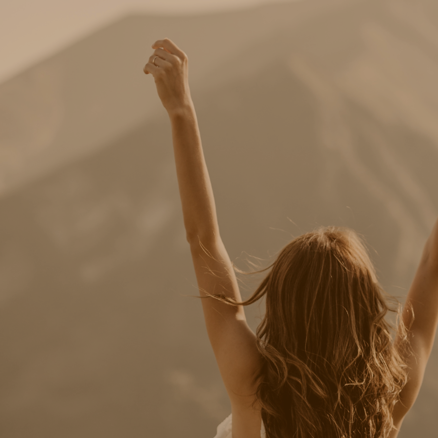 A woman with long wavy hair raising her arms in a joyful manner under soft lighting.