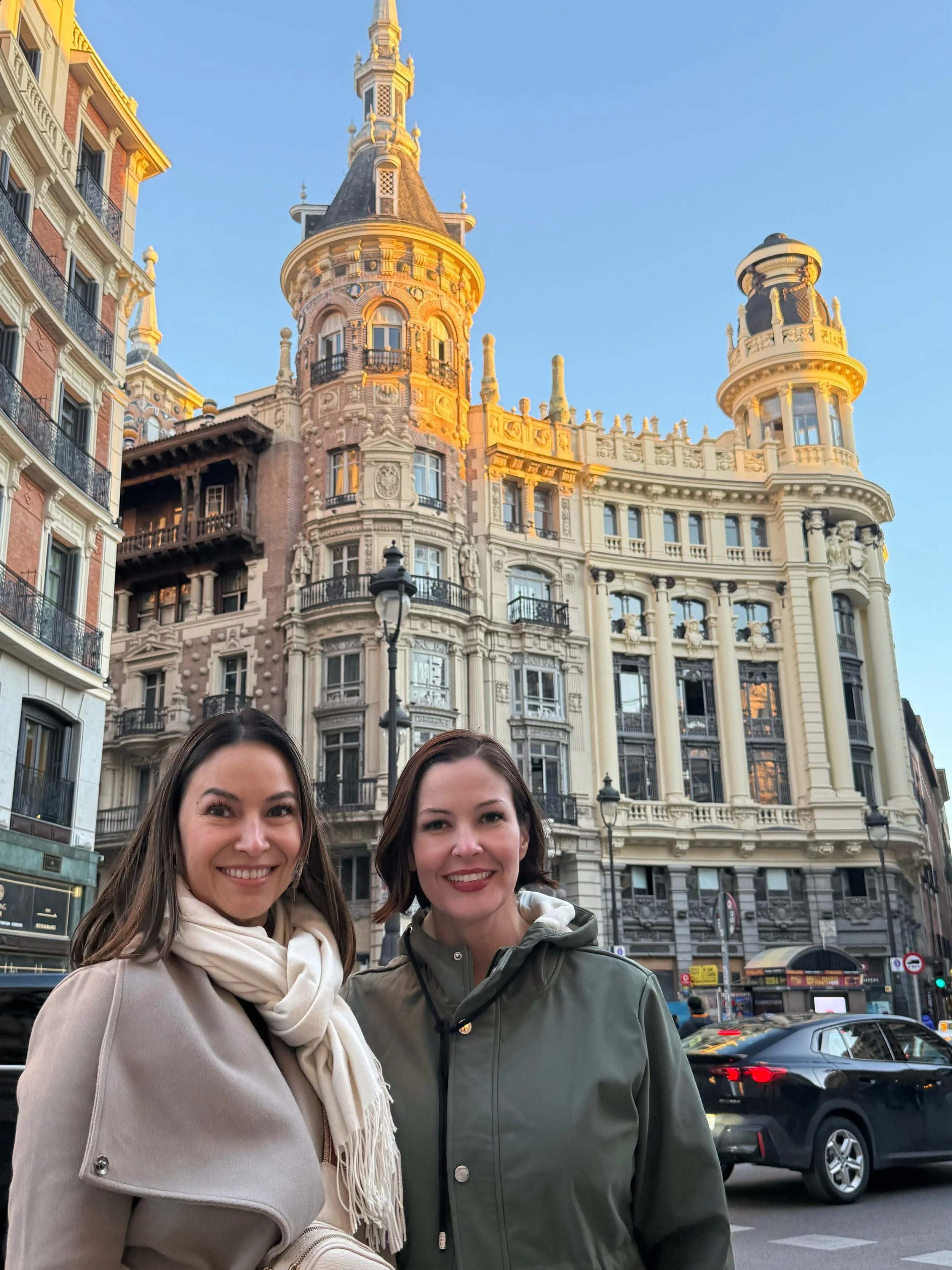 Two women with dark hair smiling in front of a historic building with ornate architecture, balconies, and towers, in an urban street scene.