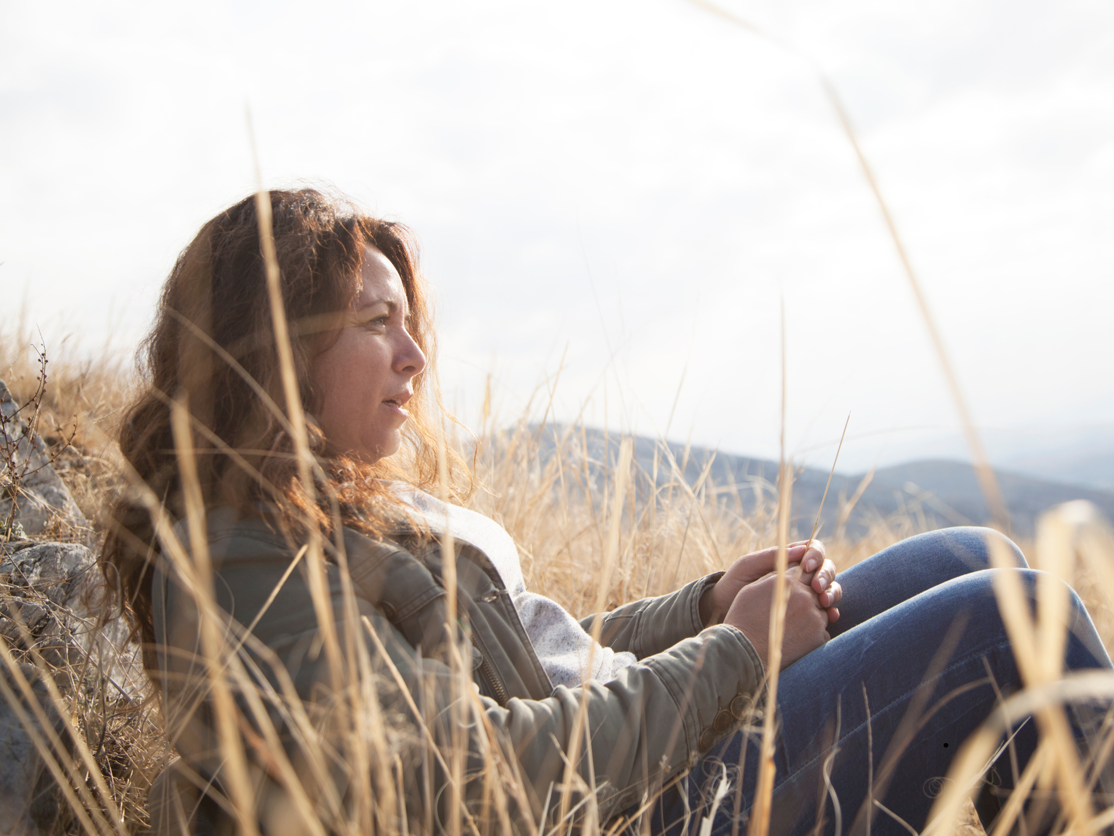 A woman with curly hair sitting outdoors in a grassy field, holding a smartphone, with mountains and a cloudy sky in the background.