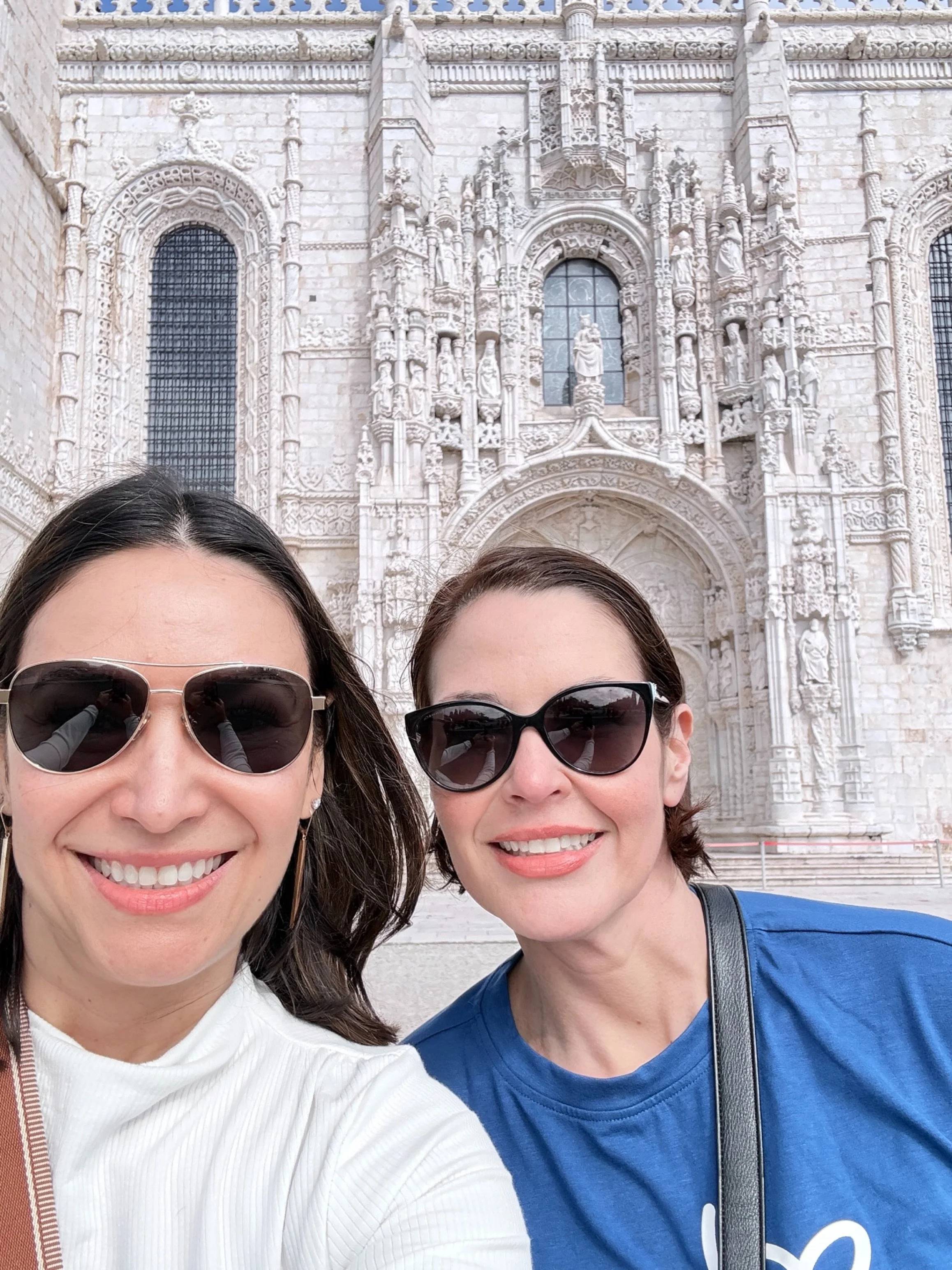 Two women smiling and wearing sunglasses in front of a historic stone building with detailed carvings and stained glass windows.