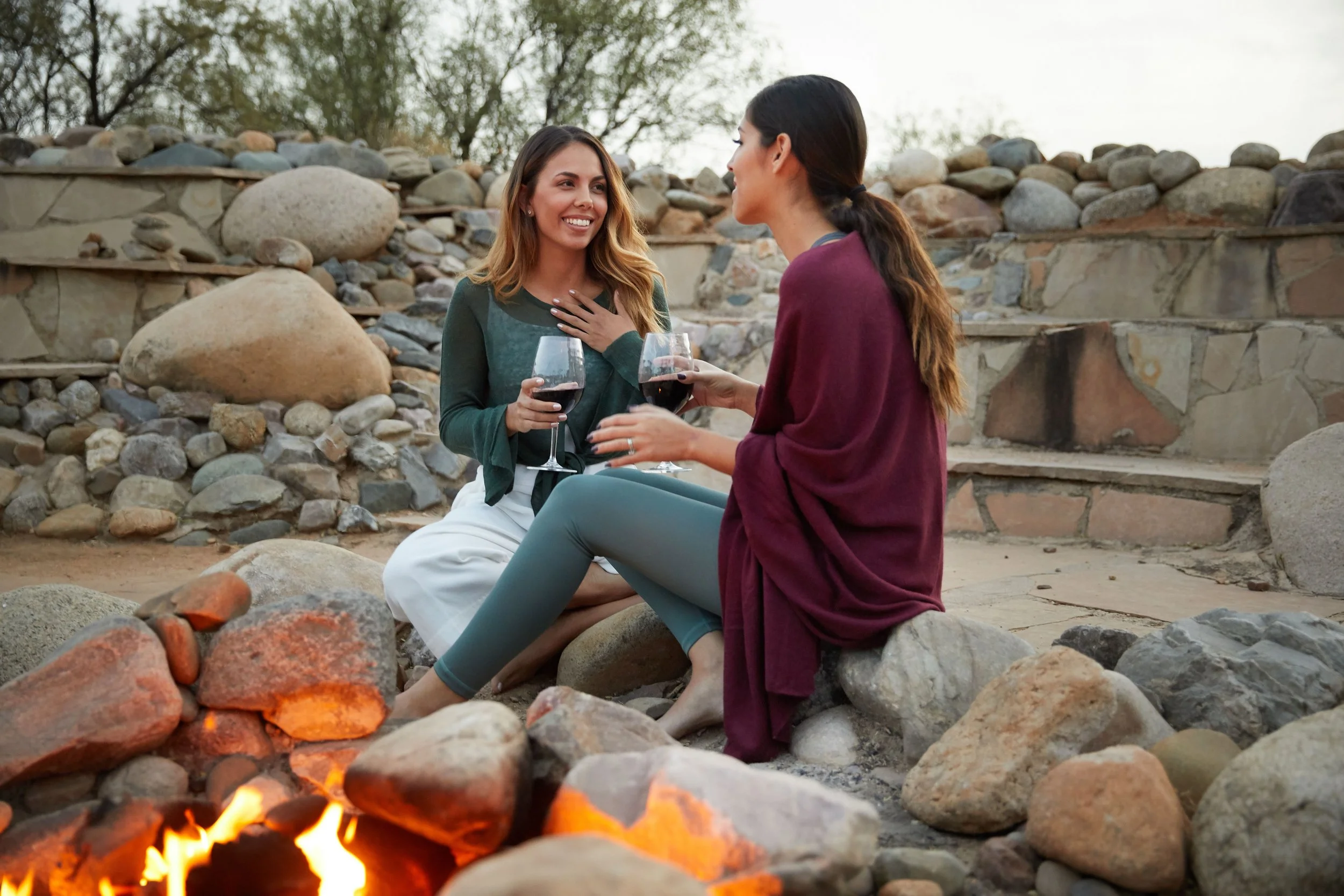 Two women sitting on rocks by a fire pit, holding glasses of red wine, chatting outdoors with stone walls and trees in the background.