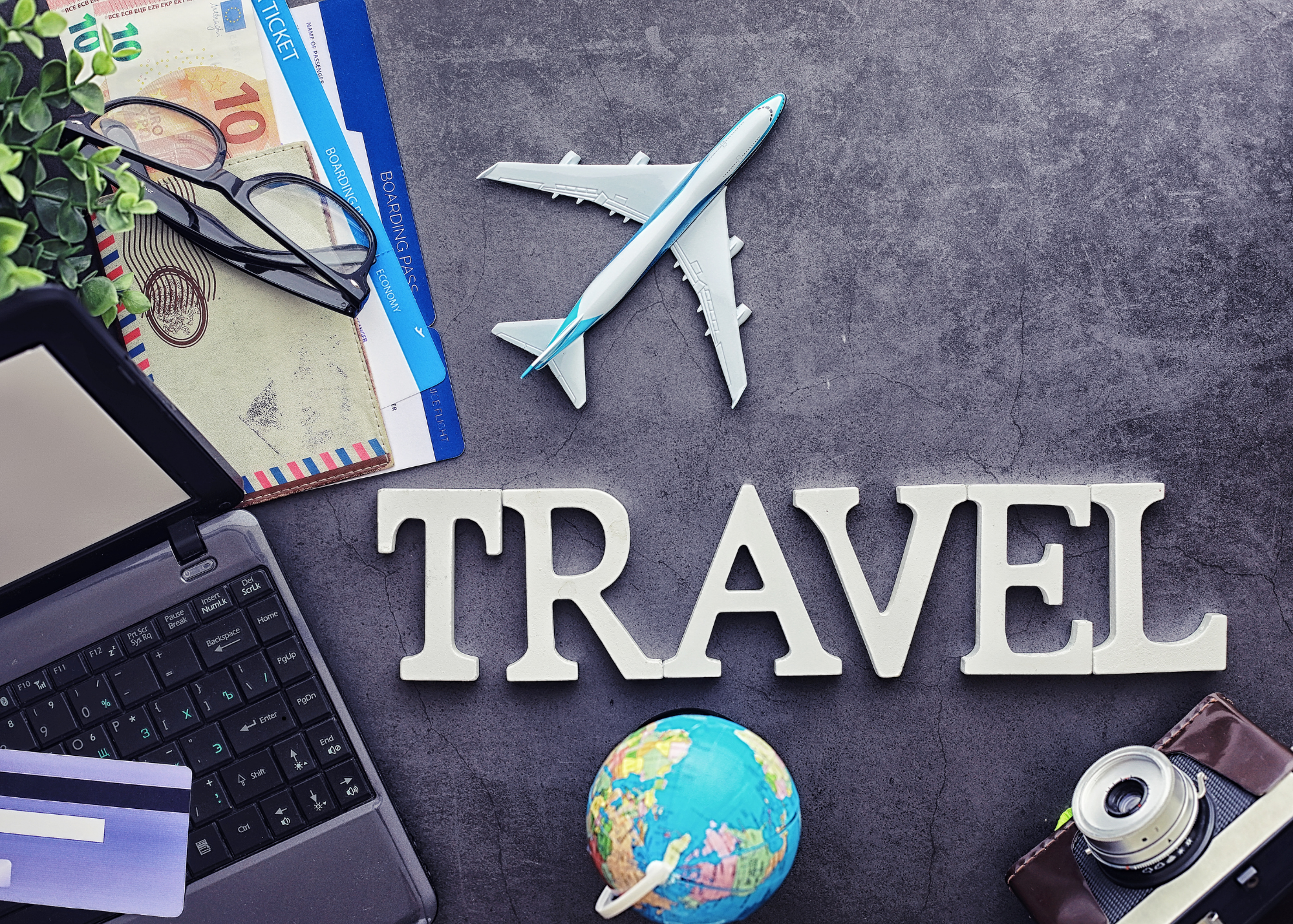 Travel-related objects on a table, including a toy airplane, an international passport, a boarding pass, Euro currency notes, glasses, a laptop keyboard, a small globe, a spool of film, and a decorative sign that reads 'TRAVEL'.