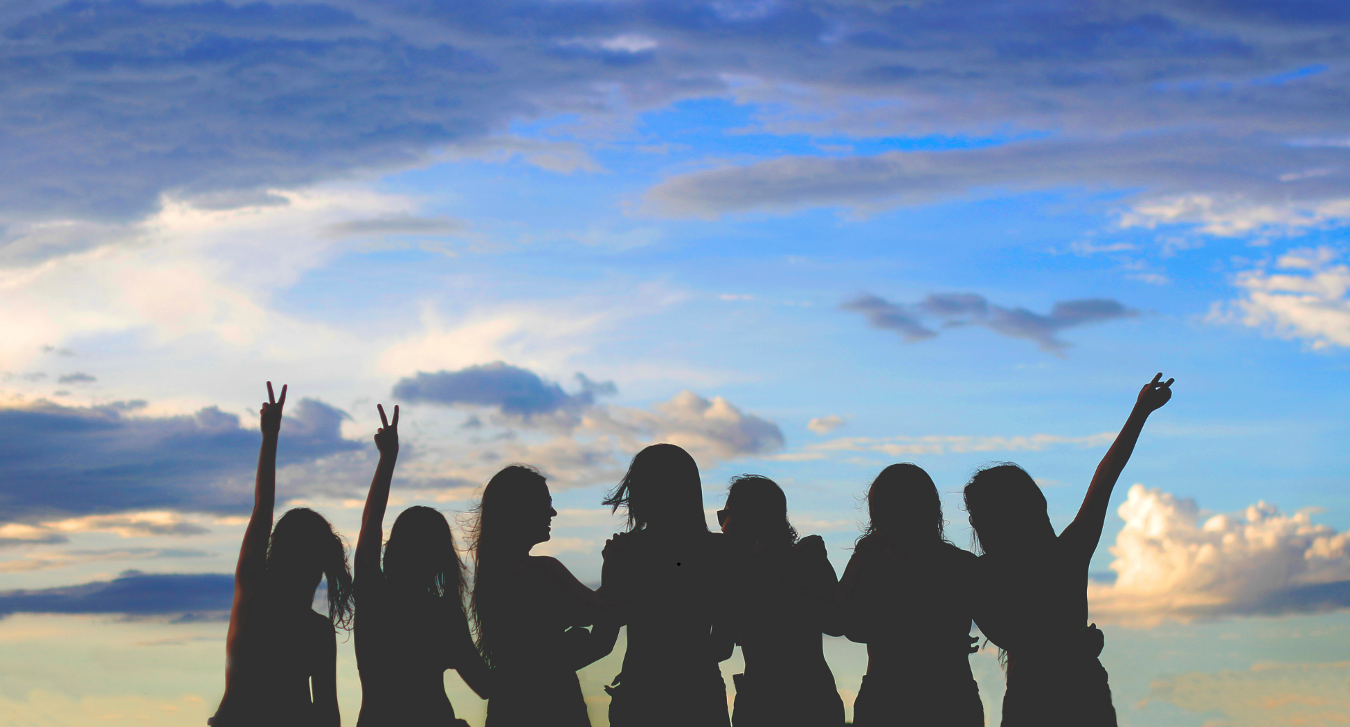 Silhouettes of seven young women standing outdoors against a partly cloudy sky, some making peace signs with their hands.