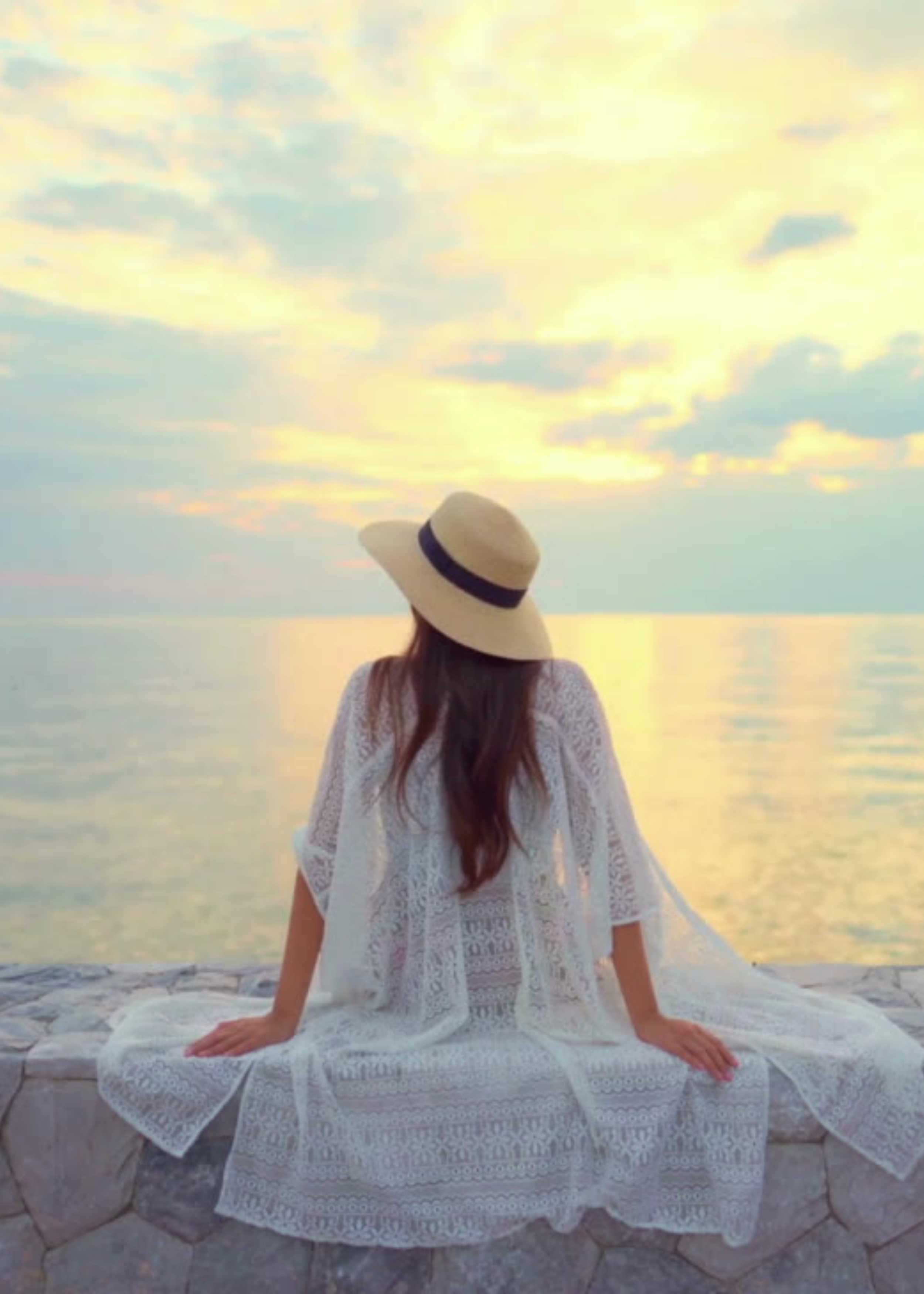 Woman wearing a white lace dress and straw hat sitting on a stone ledge by the water during sunset.