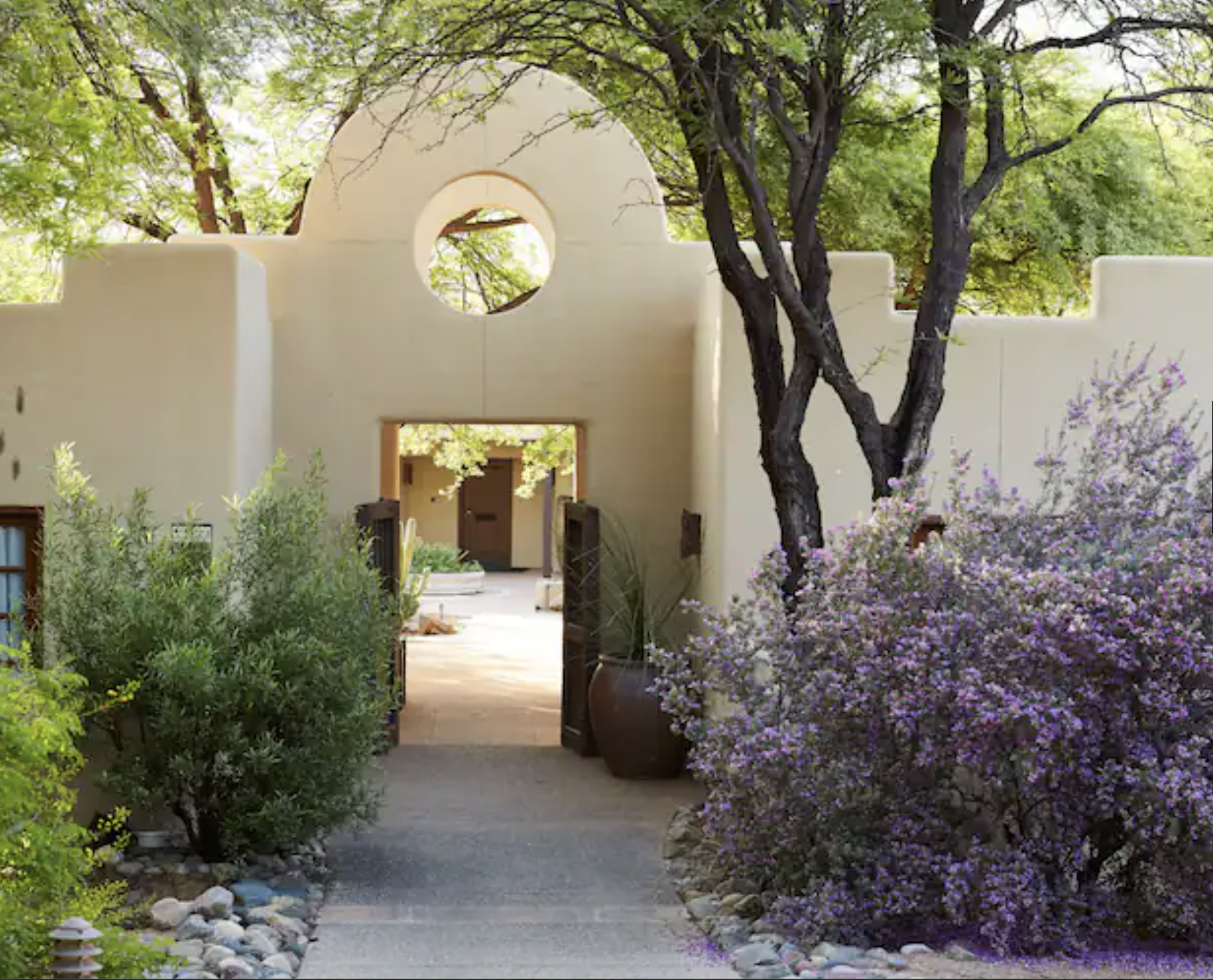 Entrance to a house with southwestern architecture, surrounded by greenery and flowering bushes, with a pathway leading to the door and a dog lying on the ground.