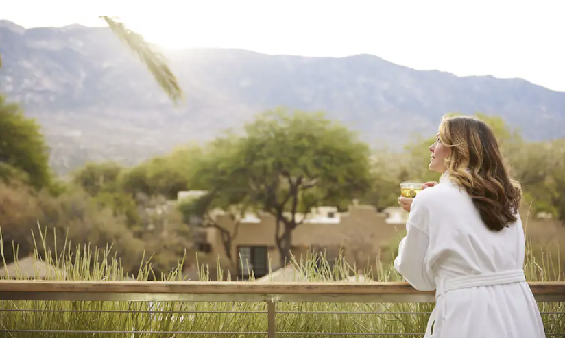 Woman in a white robe holding a glass, standing outdoors on a balcony with desert plants, trees, and mountains in the background.
