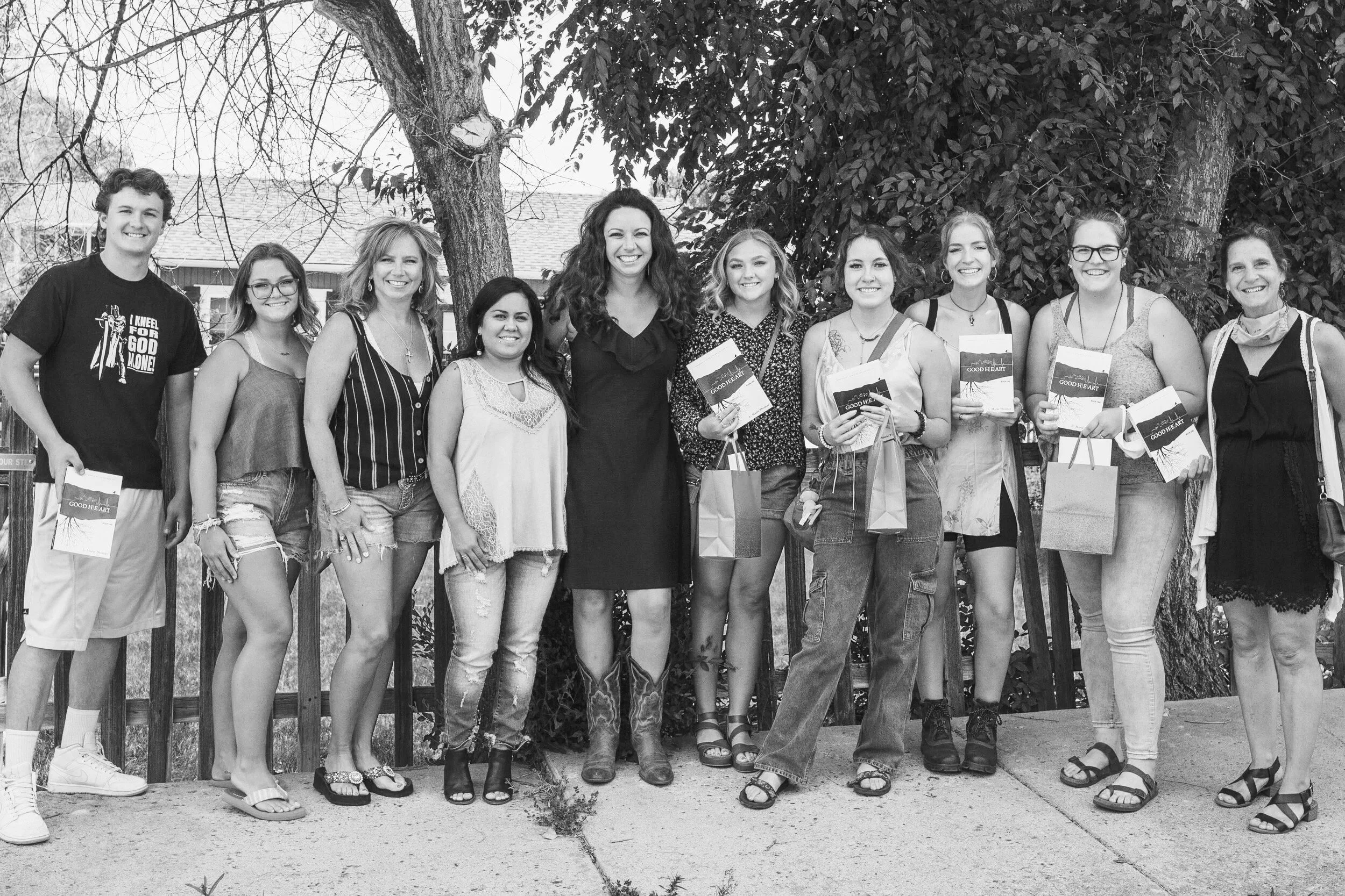 Group of women and one young man standing with L.M. Thomas—the author, mentor, and speaker—outdoors in front of trees, smiling, holding The Good Heart Series A.R.C. copy of book one and gift bags.
