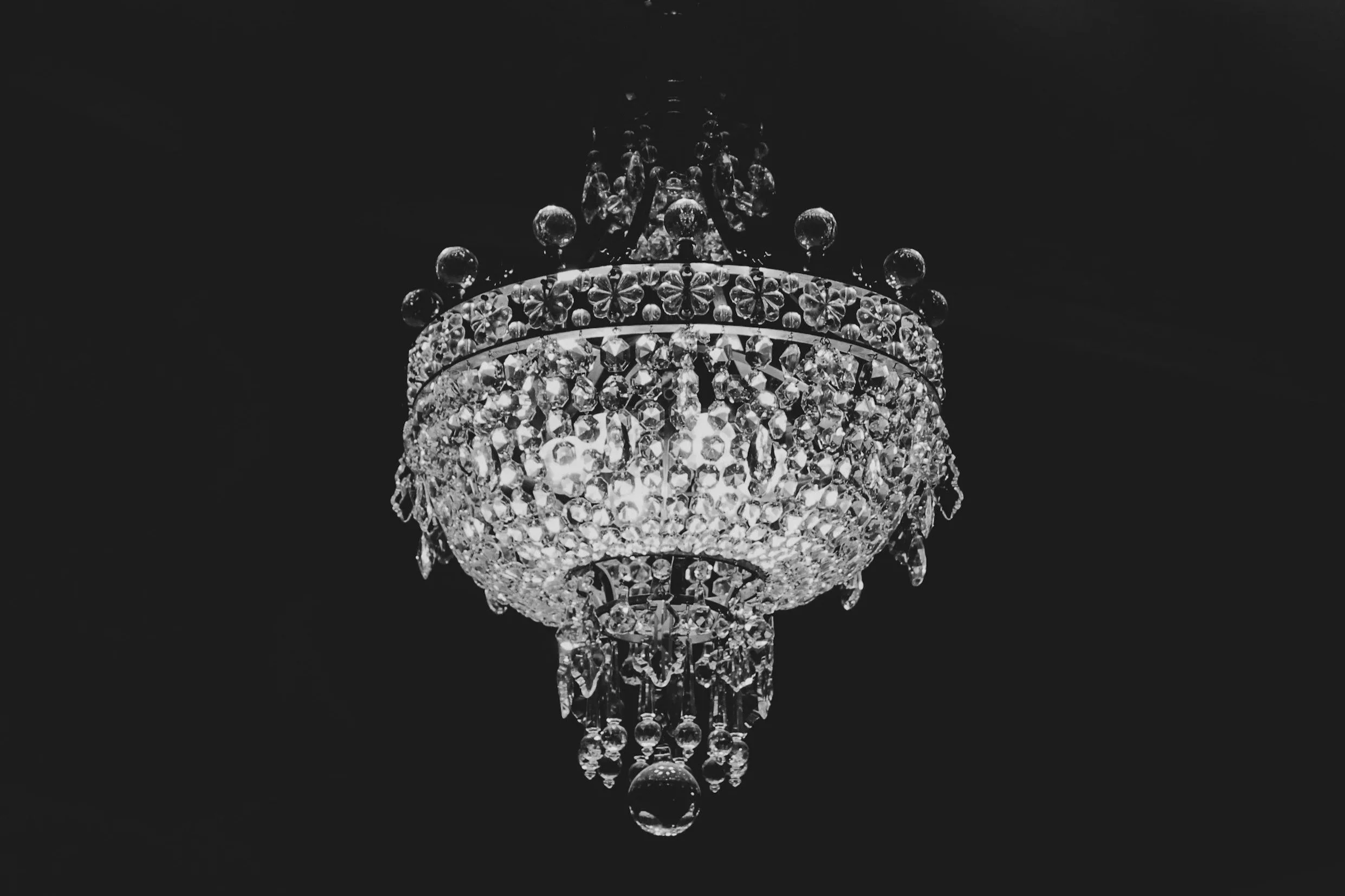 A black and white photograph of an ornate crystal chandelier hanging from a dark ceiling representing the wealth of the Bailey Family in 'The Good Heart Series' by L.M. Thomas.
