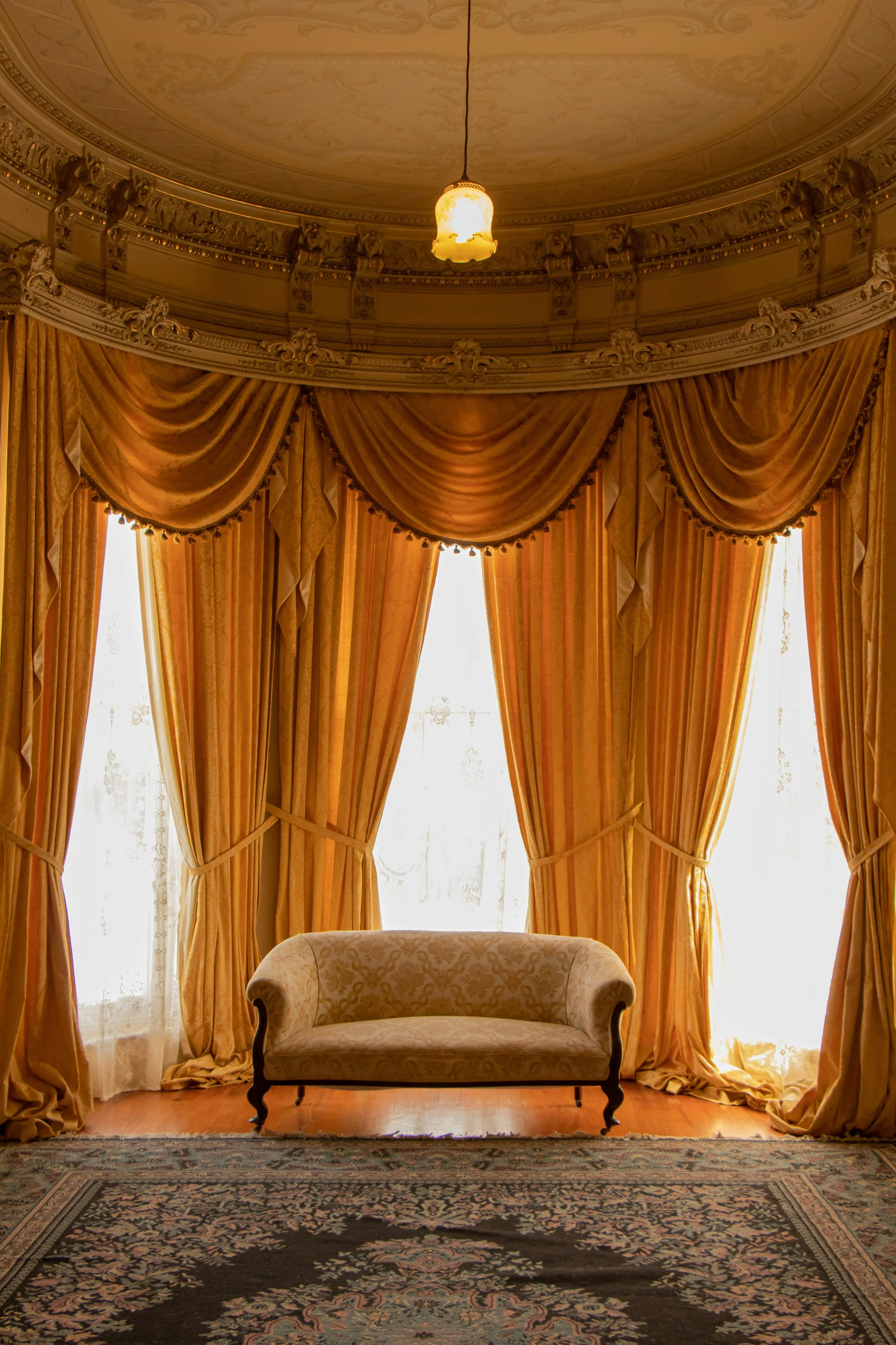 Luxurious room with large windows covered by yellow drapes, a beige upholstered sofa in front of the windows, ornate ceiling and a Persian rug on the wooden floor.