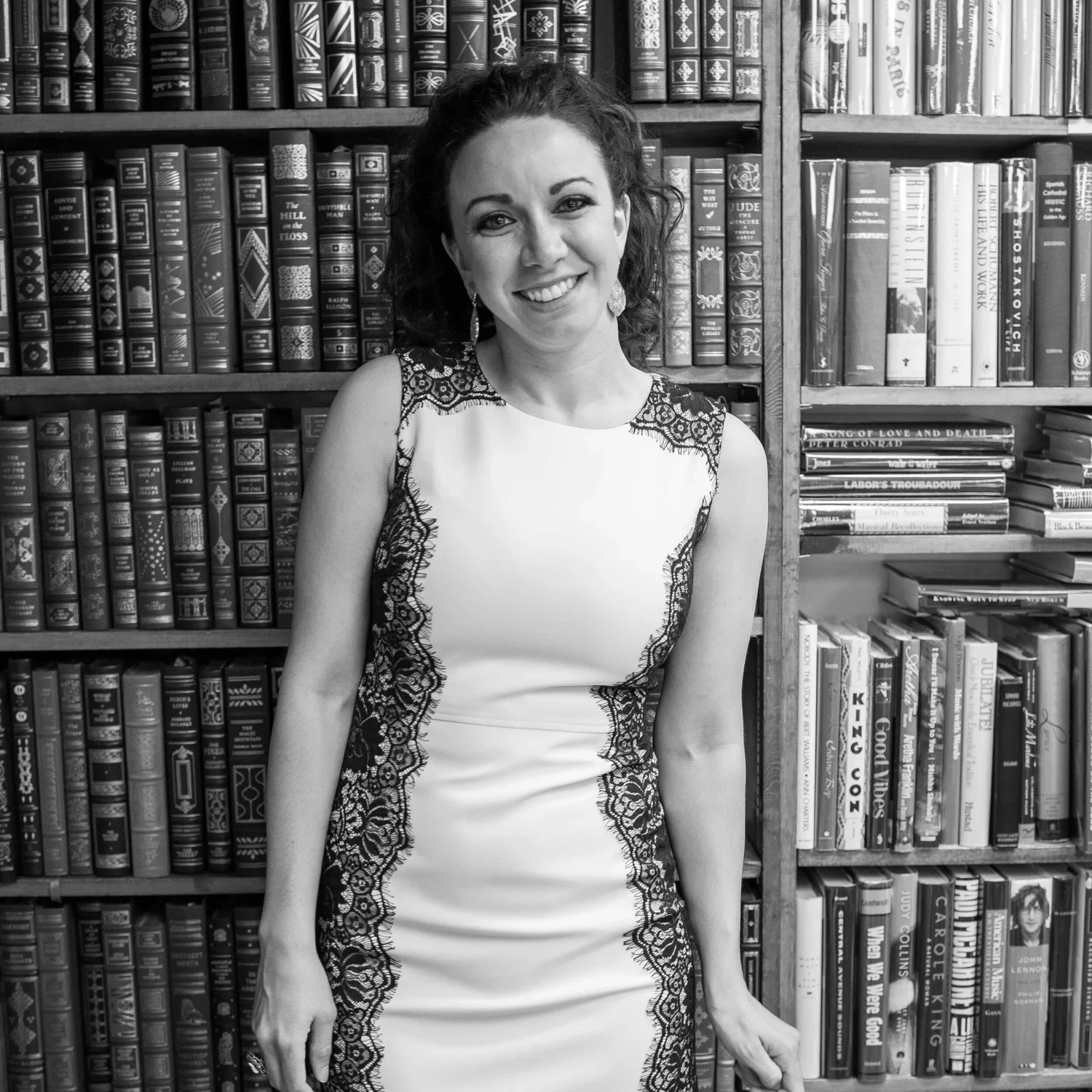 L.M. Thomas—author, speaker and mentor—in a sleeveless dress with lace details standing in front of bookshelves filled with books like 'The Good Heart Series".