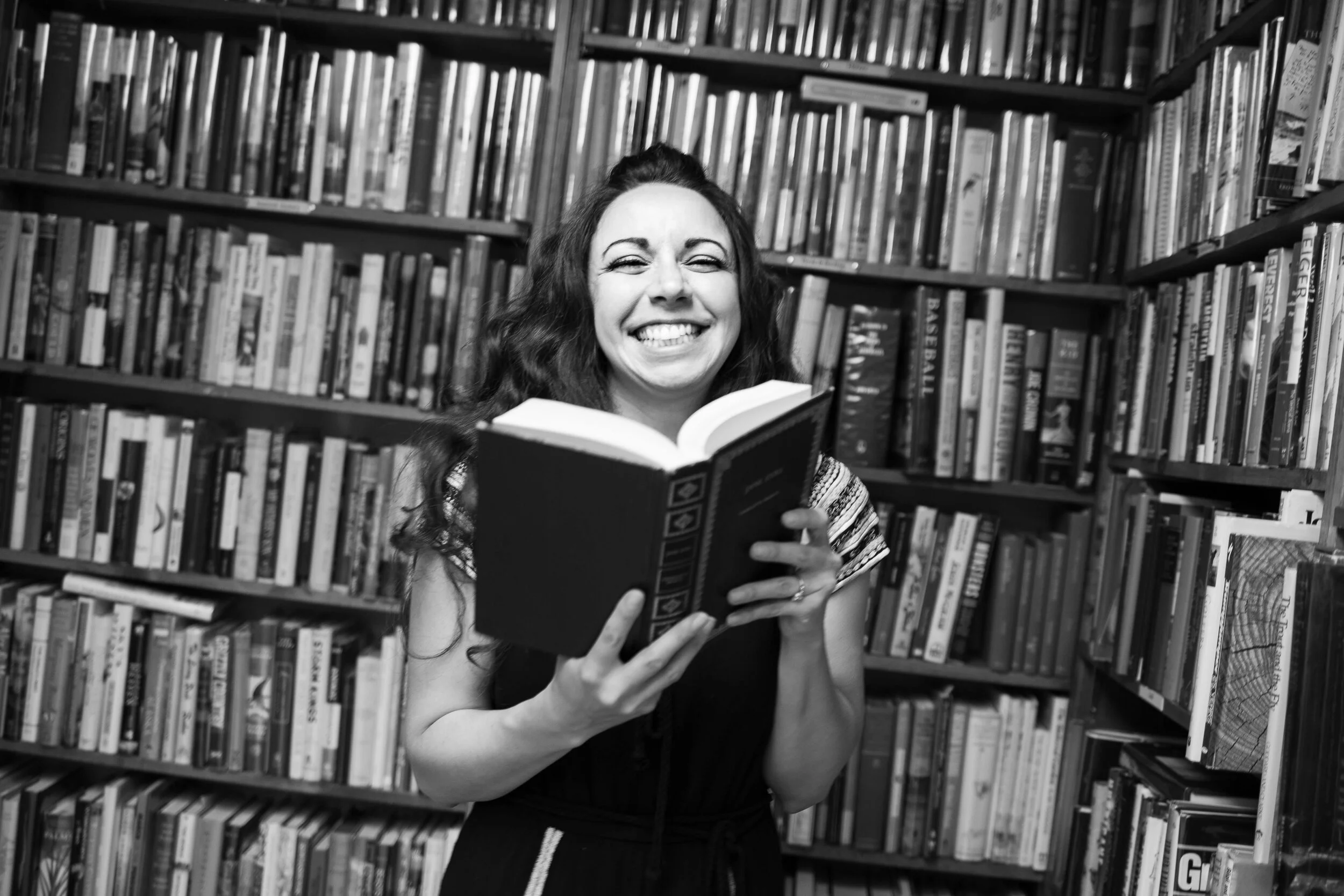 L.M. Thomas sincerely smiling while holding a book in a library surrounded by bookshelves filled with books like The Good Heart Series.