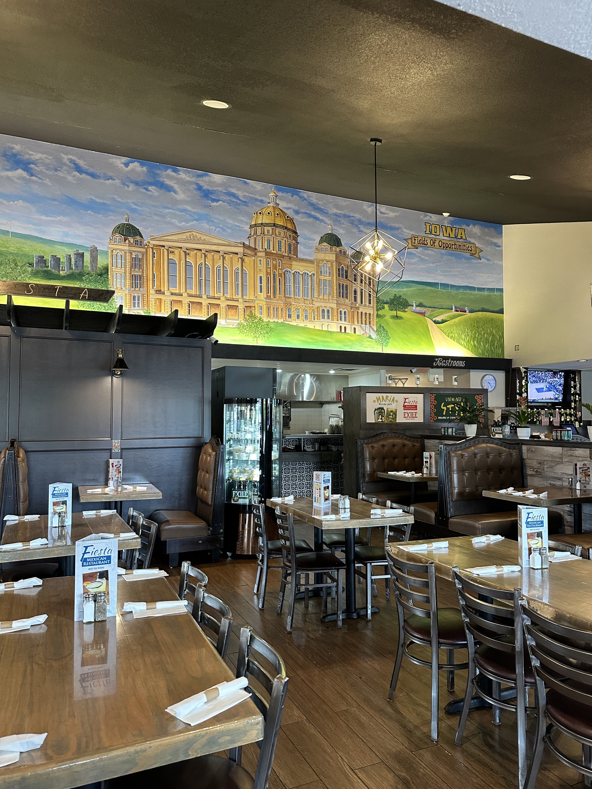 Interior of a restaurant with wooden tables, chairs, and booths, with a mural on the wall featuring a large building and green landscape, and a chandelier hanging from the ceiling.
