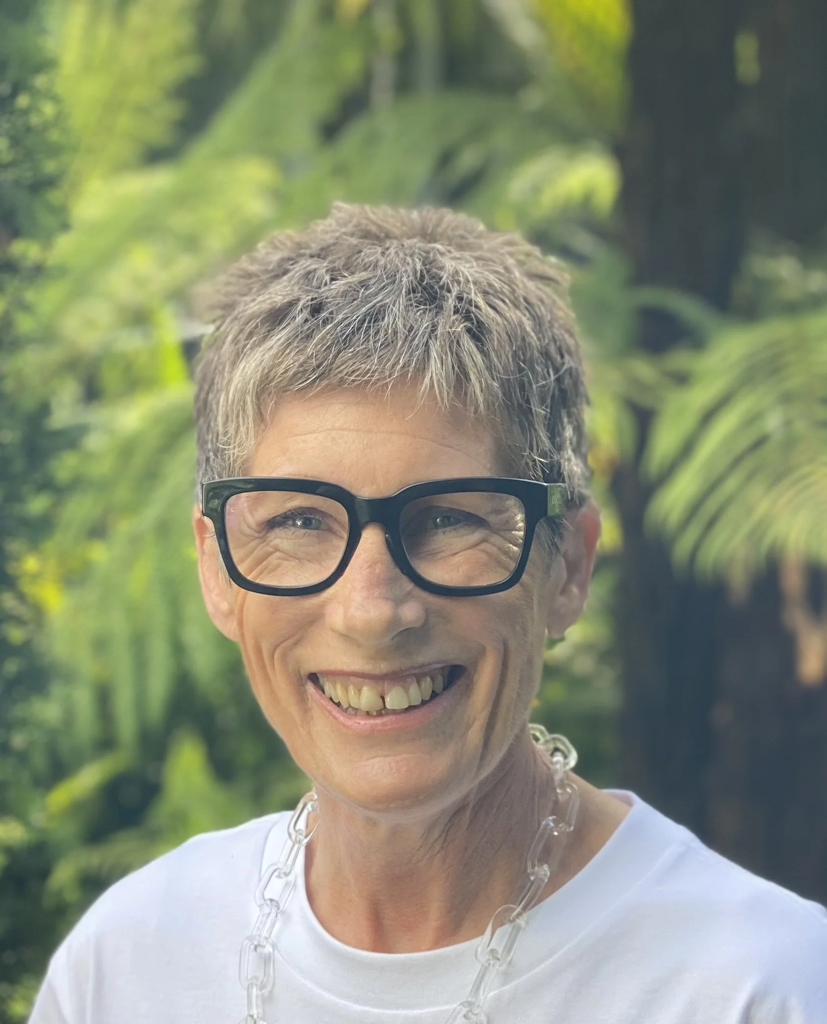 A woman with short, curly gray hair wearing glasses, a white shirt, and a necklace, smiling outdoors with green foliage in the background.