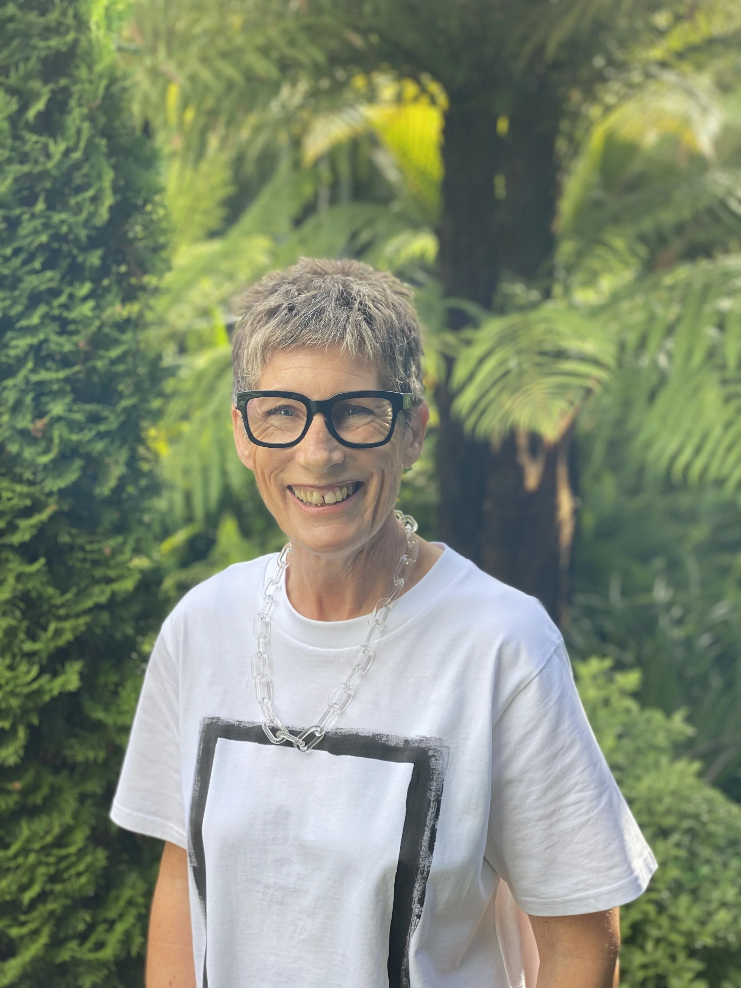 A smiling older woman with short gray hair, wearing glasses, a white T-shirt with a black rectangular design, a chunky clear necklace, standing outdoors with lush green foliage in the background.