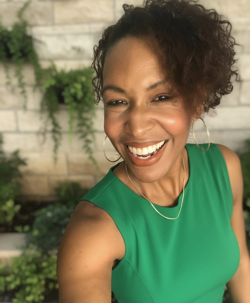 A woman with short curly hair wearing a sleeveless green top, smiling outdoors with a brick wall and green plants in the background.
