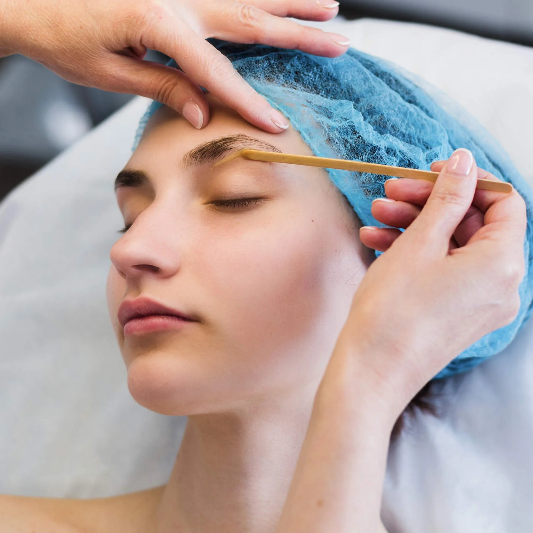 A woman receiving a cosmetic eyebrow waxing, lying down with eyes closed, wearing a blue head cap, while a practitioner applies a wax  with a small wooden applicator.