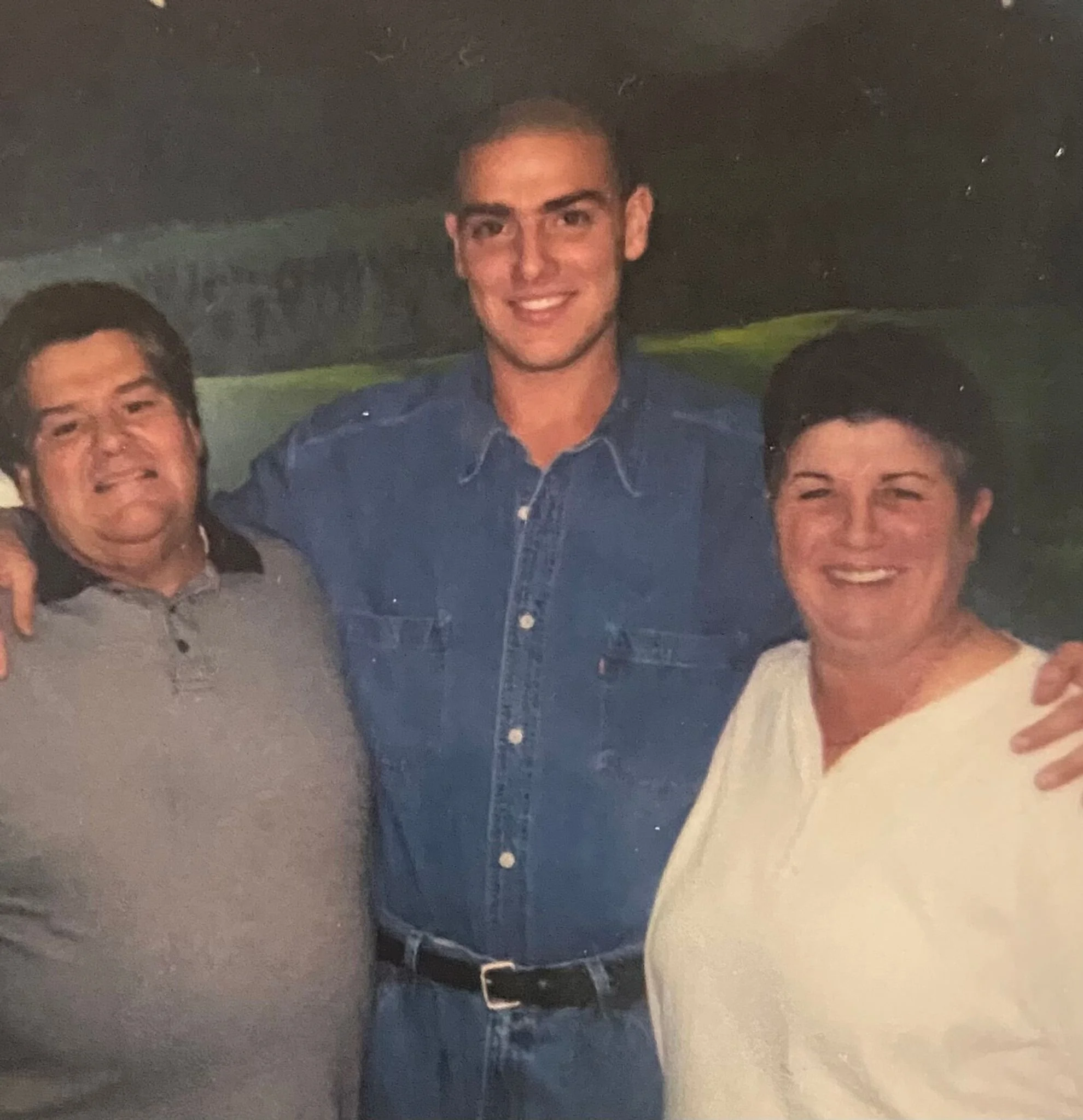 Three people smiling and standing close together outdoors, with a dark background and mountains in the distance. The person in the middle is wearing a denim shirt, while the person on the left is wearing a light gray polo shirt and the person on the right is wearing a white blouse.