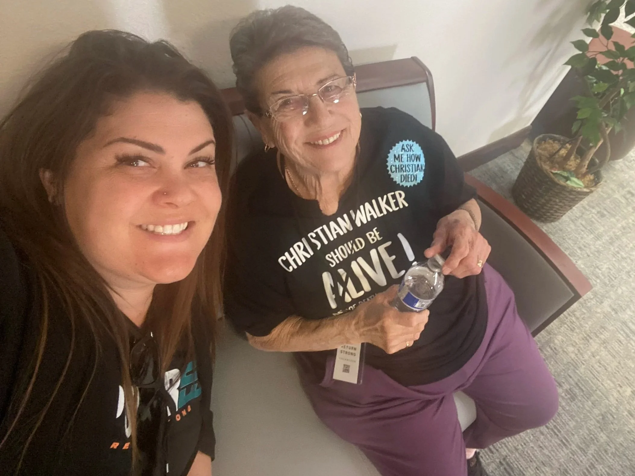 Two women sitting on a bench, smiling for a selfie. The older woman is holding a water bottle and wearing a black shirt with white text, purple pants, and glasses. The younger woman has long brown hair and is wearing a dark shirt with colorful writing.