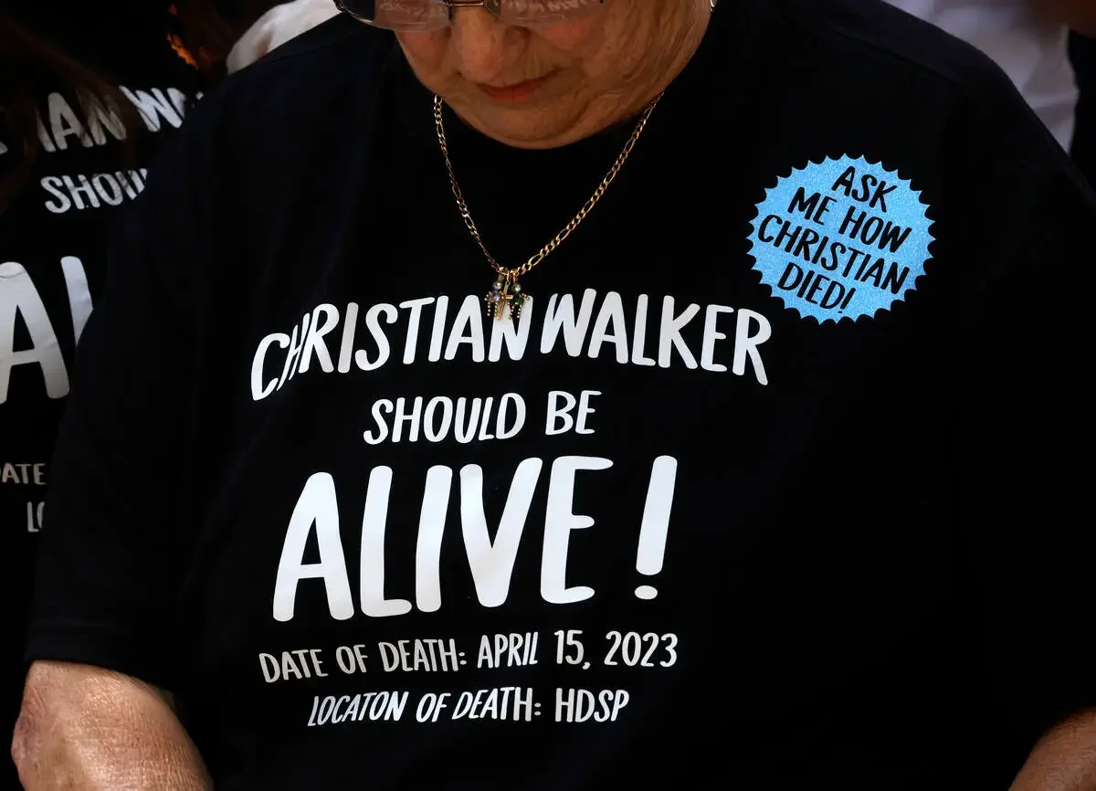 Close-up of a person wearing a black t-shirt with white text that advocates for Christian Walker, stating he should be alive, with details about his death on April 15, 2023, in HDSP. A blue sticker on the shirt asks, "Ask me how Christian died!" The person also wears a gold chain with a cross pendant.