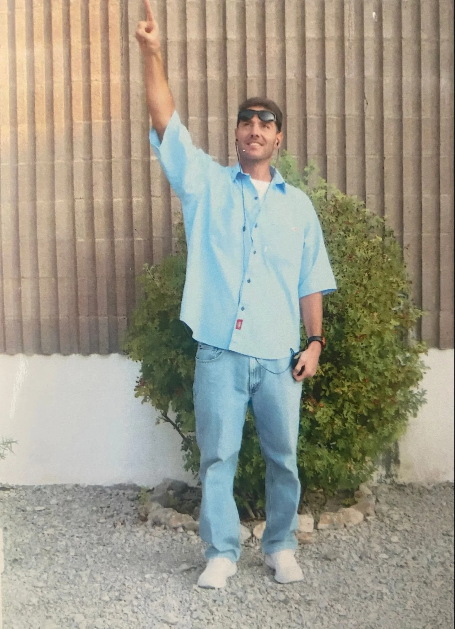 A man standing outdoors in front of a wooden fence and a bush, wearing a light blue shirt, jeans, sunglasses, and a smartwatch, smiling and pointing upward.
